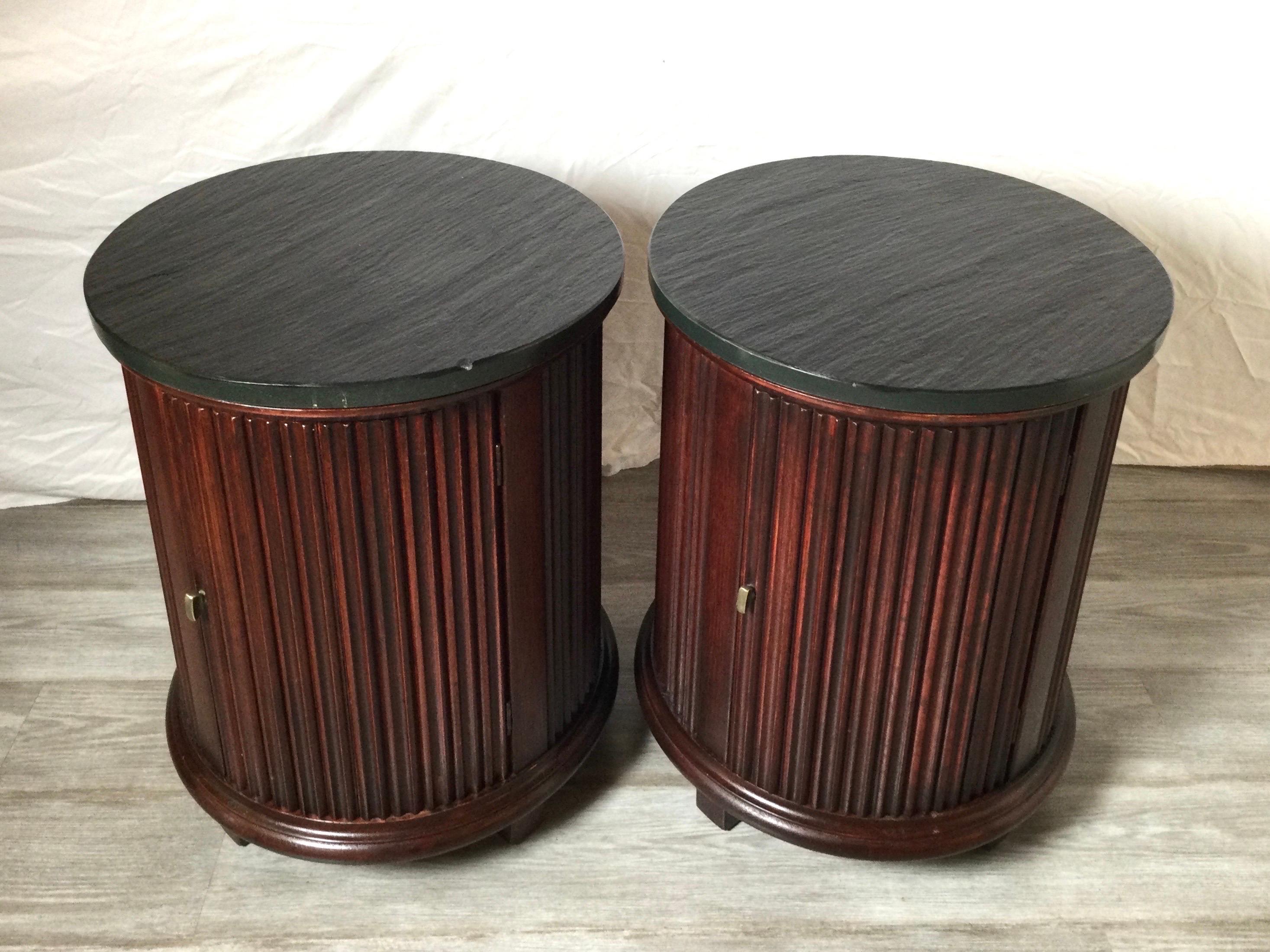 A stylish pair of round storage side tables with natural slate tops. The textured slate tops in dark grey with ribbed mahogany curved sides with a well designed concealed door that opens to reveal shelved storage.