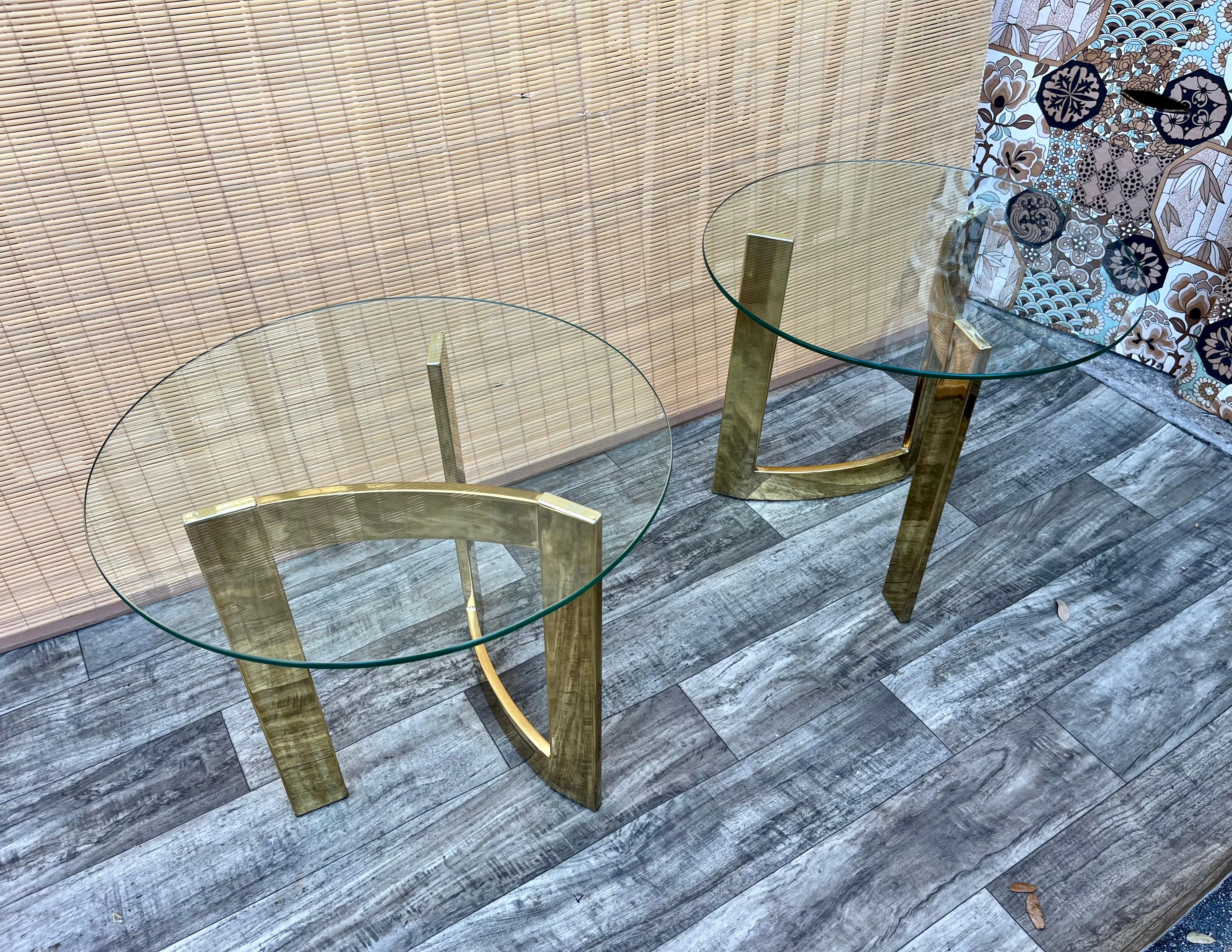 A Pair of Vintage Mid Century Modern / Hollywood Regency Brass tables in The Milo Baughman Style. Circa 1970s  Feature arched triangular shaped brass plated bases with removable round glass tops. 
In Excellent Original Condition With Minor Signs of