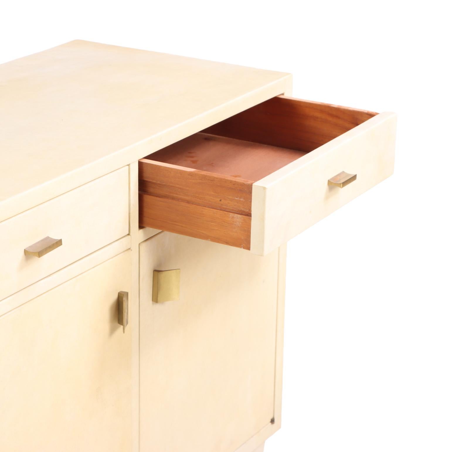 A refined pair of mid-century modern cabinets, circa 1960, each fully wrapped in natural parchment with a soft, matte sheen that subtly enhances their clean, architectural lines. The minimalist form is balanced by understated brass hardware, lending