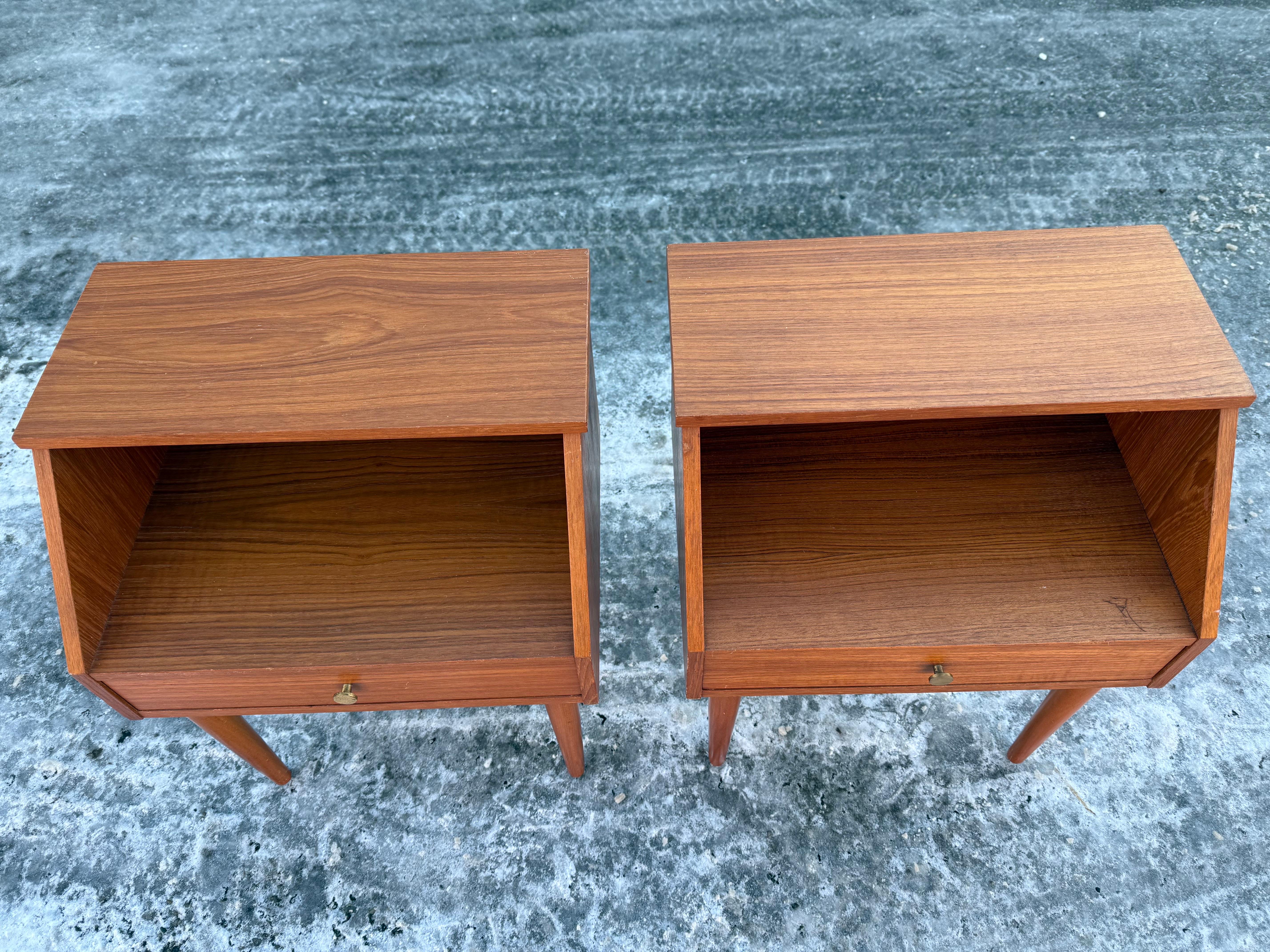 A stunning pair of mid-century modern nightstands from the 1960s, crafted in rich teak. Featuring a sleek, minimalist design with a spacious open shelf and a single drawer with a brass pull, these elegant pieces rest on tapered legs, embodying the