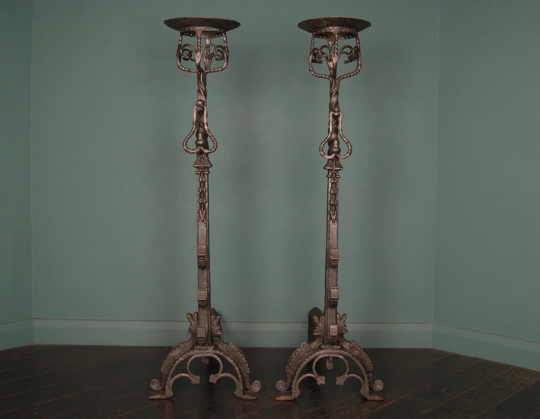 A monumental pair of Neo-Gothic wrought-iron fireplace andirons or fire dogs, extensively adorned with fine blacksmith detail. The standards incorporate ornate spit-hooks, acorn front-handles and cresset finials with inter-twinned serpent and dragon