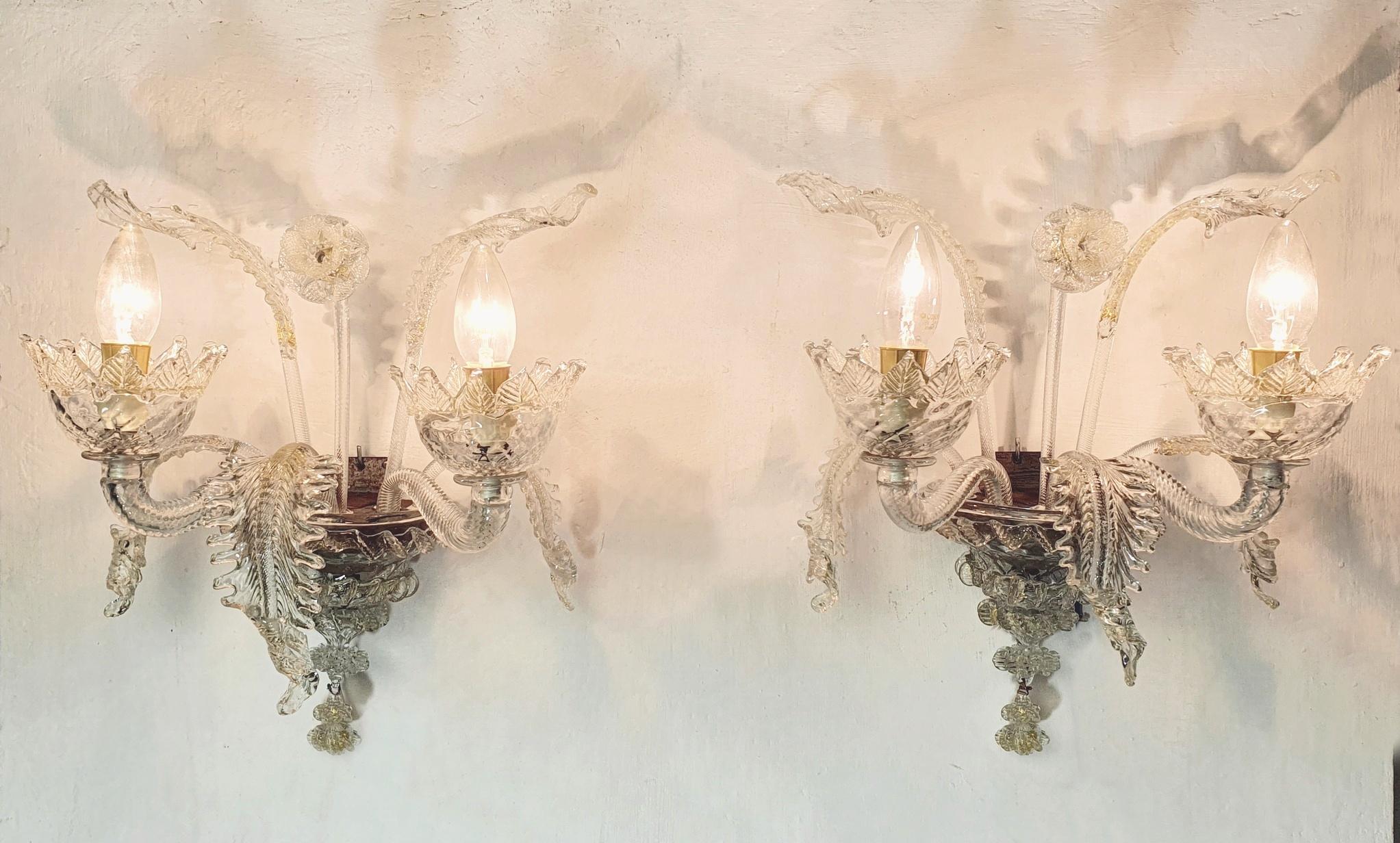 A nice pair of classic Murano sconces in Venetian style in clear glass with gold specks giving the light a nice shimmer.  There is a flower in the middle of each sconce as well as five curled leaves. The sconces surrounding the lightbulb have a wide