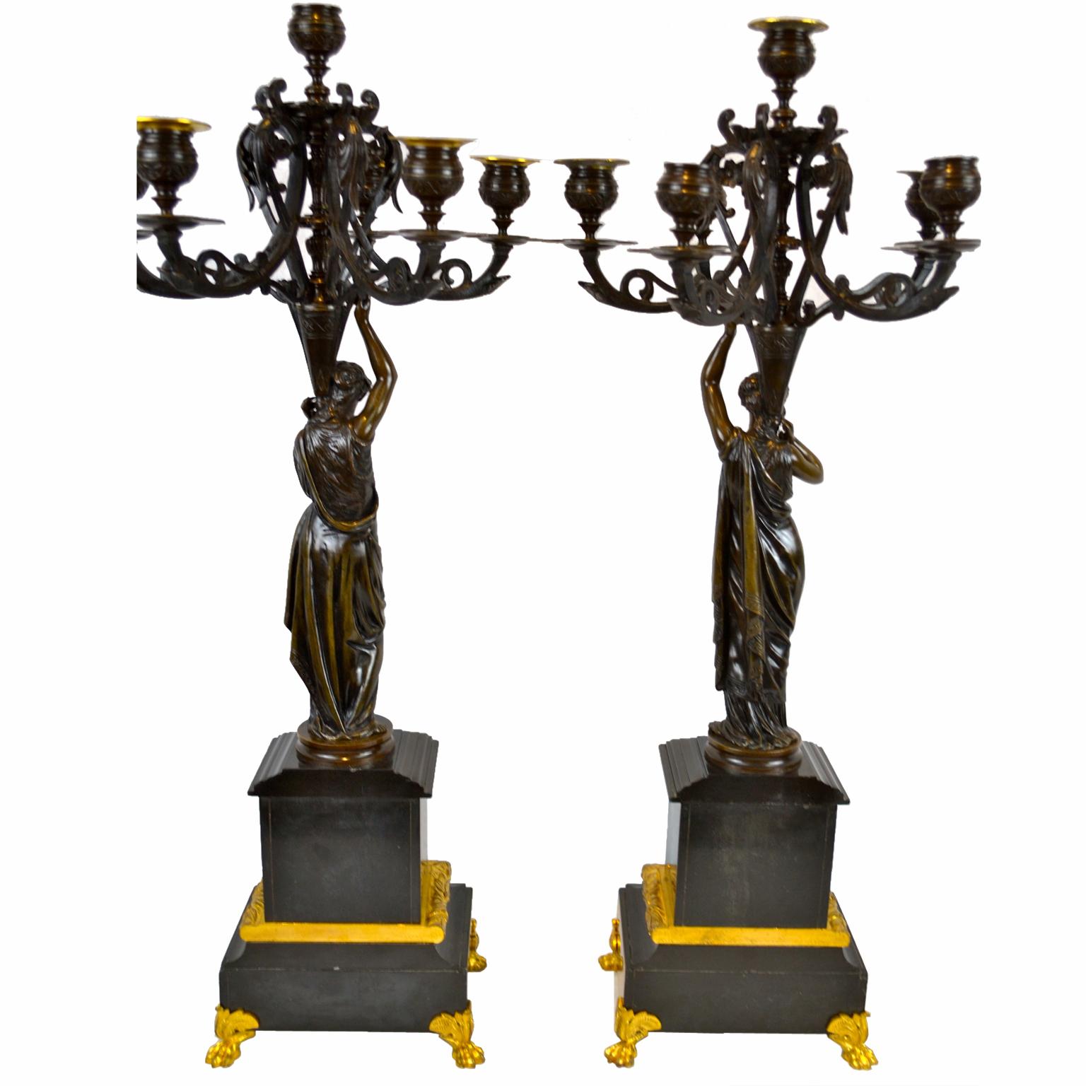 Very well cast models of classical maidens standing on decorated stepped rectangular black marble bases, the upper base mounted with an Etruscan style frieze; the bases have gilt bronze trim and gilt bronze lions feet. Each maiden holds aloft a