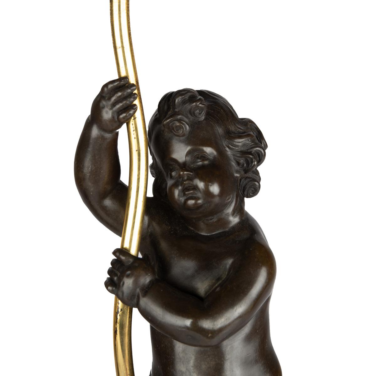 Pareja de lámparas de sobremesa Napoleón III de bronce y ormolina, cada una en forma de putto de bronce que sostiene la base de una lámpara de una sola luz, de mano izquierda y derecha, asentadas sobre una base cilíndrica de ormolina decorada con