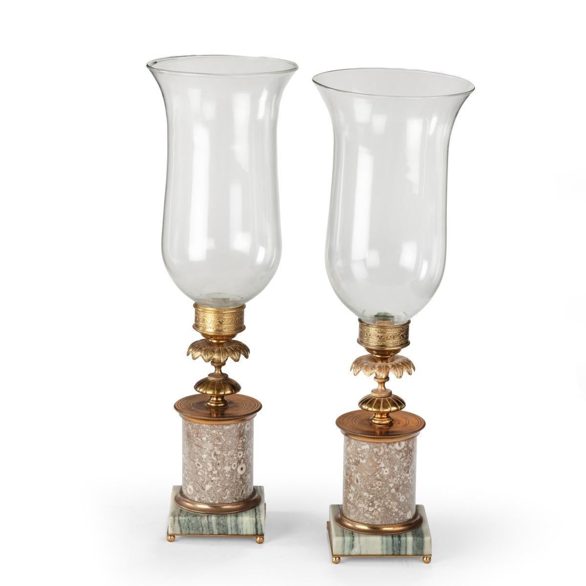 A pair of decorative storm lamps, each with a candle holder enclosed in a clear glass shade set on a turned gilt-brass support with a collar of petals and a cylindrical marble column, the rectangular base of striped marble.  English, early 19th
