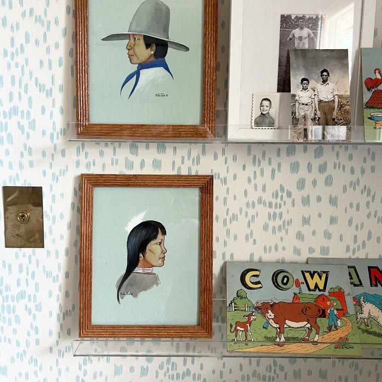 A framed pair of a Native American man in woman. These paintings are by Oklahoma artist Jon Tiger. They were created in 1991 and feature a side profile portrait painting of the two. The woman wears a traditional native dress with white and pink