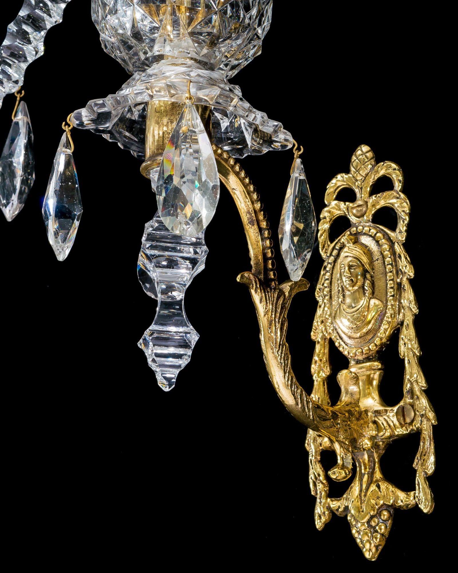 The rare pair of Neoclassical wall lights, are suspended from ornate back plates with roman mofits, mounting a diamond cut container issuing three facet cut arms, supporting van-dyke canopies and candle nozzles, central notch cut spire, with a