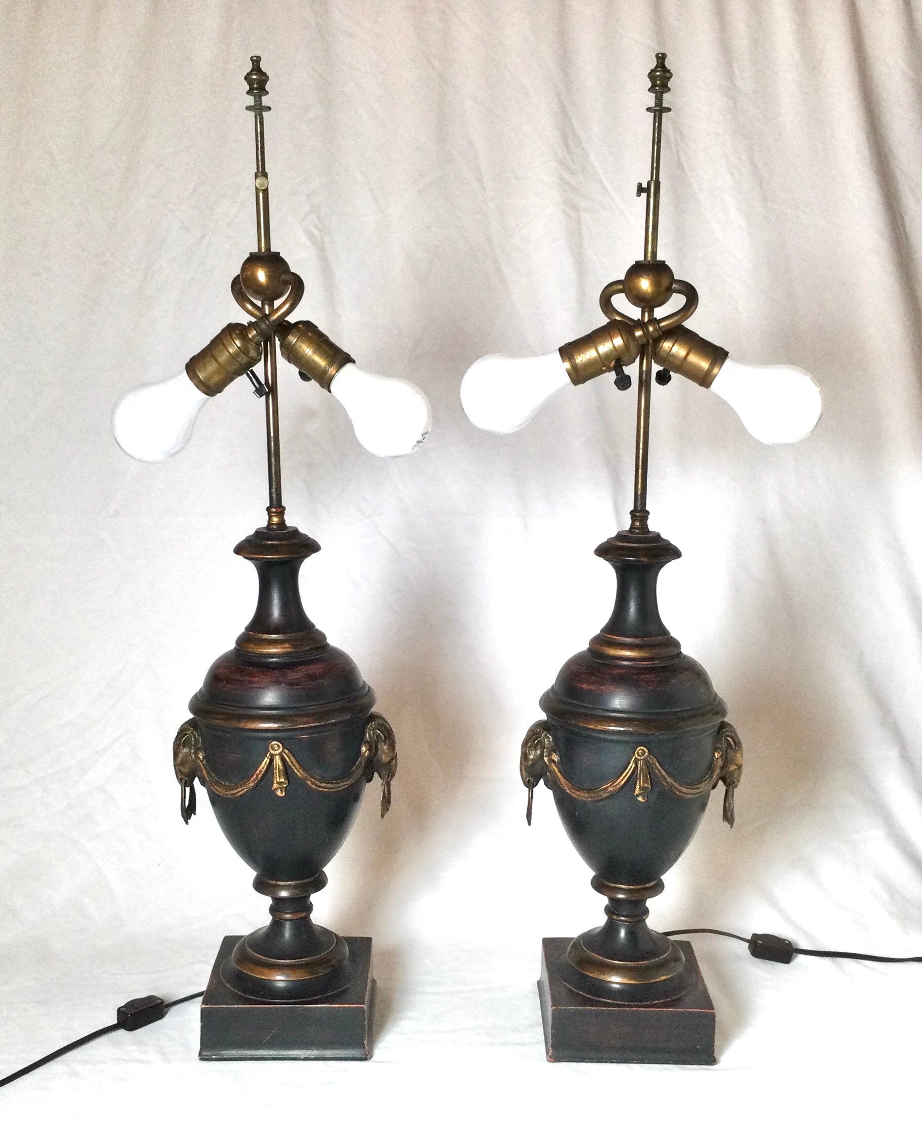 A handsome pair of urn form lamps with a painted and patinated finish. The urns with rams head handles and draped decoration on the front and back. The shades are for photographic purposes only and not included with the lamps. 29 inches tall with a