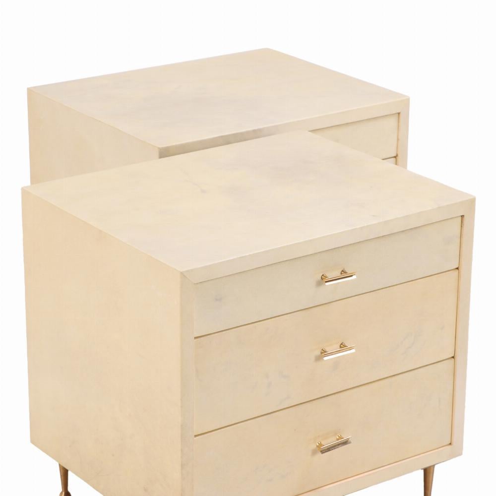 Una sofisticata coppia di armadietti o comodini a tre cassetti, circa 1950, rivestiti in pergamino naturale dai toni ricchi. Le forme pulite e rettilinee sono completate da una ferramenta in ottone minimale e da gambe corte e tornite, che permettono