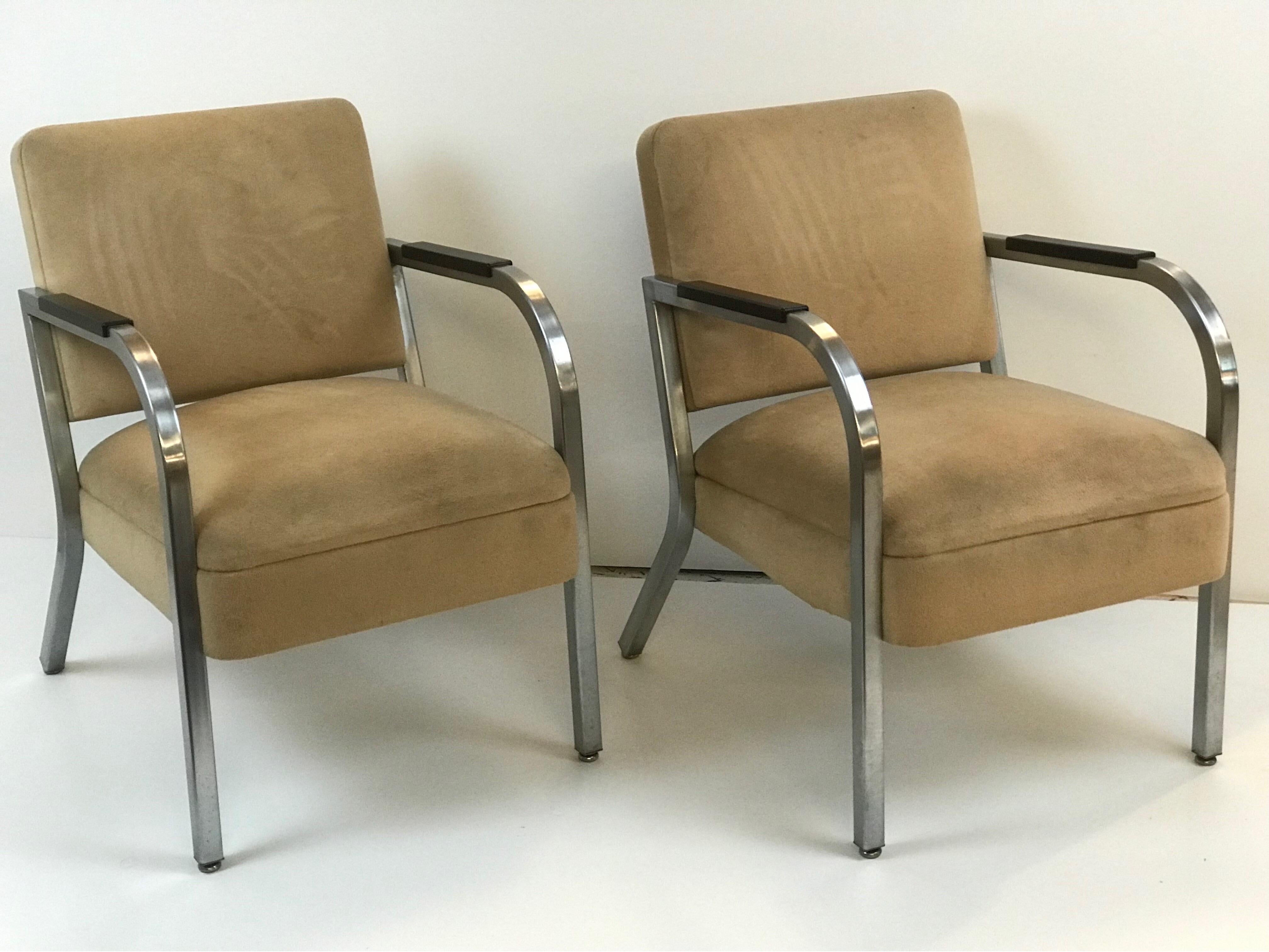 A pair of 1960's petite bent square steel tubing club chairs with black melamine arms set on medium luster polished stainless steel exposed framing with newer 2000's era tan Ultrasuede upholstery. 
Unknown origins as they are untagged and don't pop