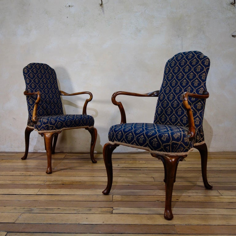 A striking pair of Queen Anne style walnut open armchairs. Displaying elegantly shaped Shephard crook arms, raised on cabriole front legs, with gracefully swept rear legs - both terminating on pad feet. The chairs are upholstered in a fleur-de-lis