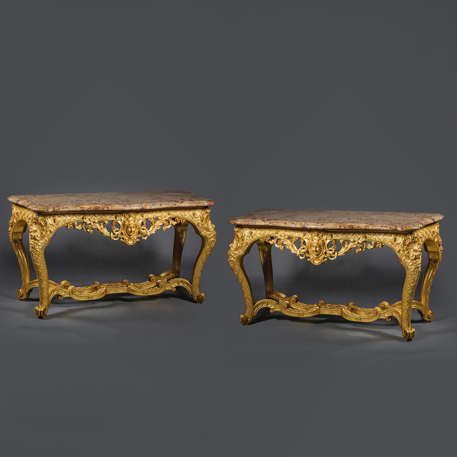 Paire de consoles en bois doré sculpté de style Régence. 

Ces consoles palatiales sont finement sculptées, la frise étant centrée par un masque entre des plumes de feuilles d'acanthe percées. Les lourds plateaux en marbre Brêche d'Alep sont