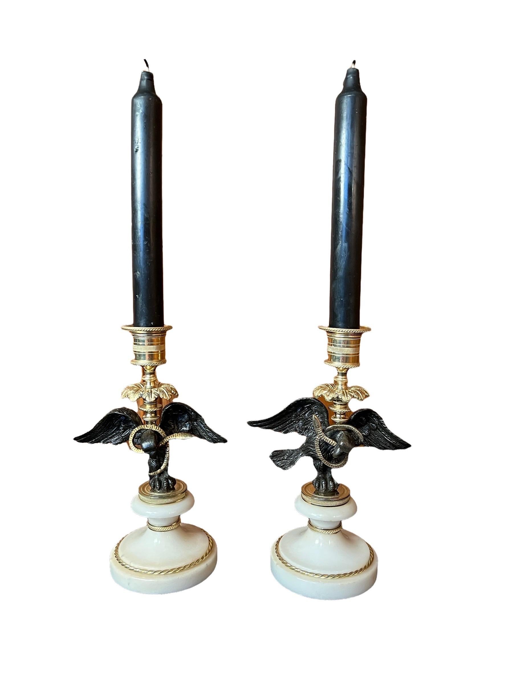 A Pair of Regency Bronze, Gilt-bronze & Marble Candle sticks. Gilt-bronze candle cup above black paginated  bronze eagles. The eagles clutching serpents, mounted on round white marble bases decorated with gilt-bronze rings. 