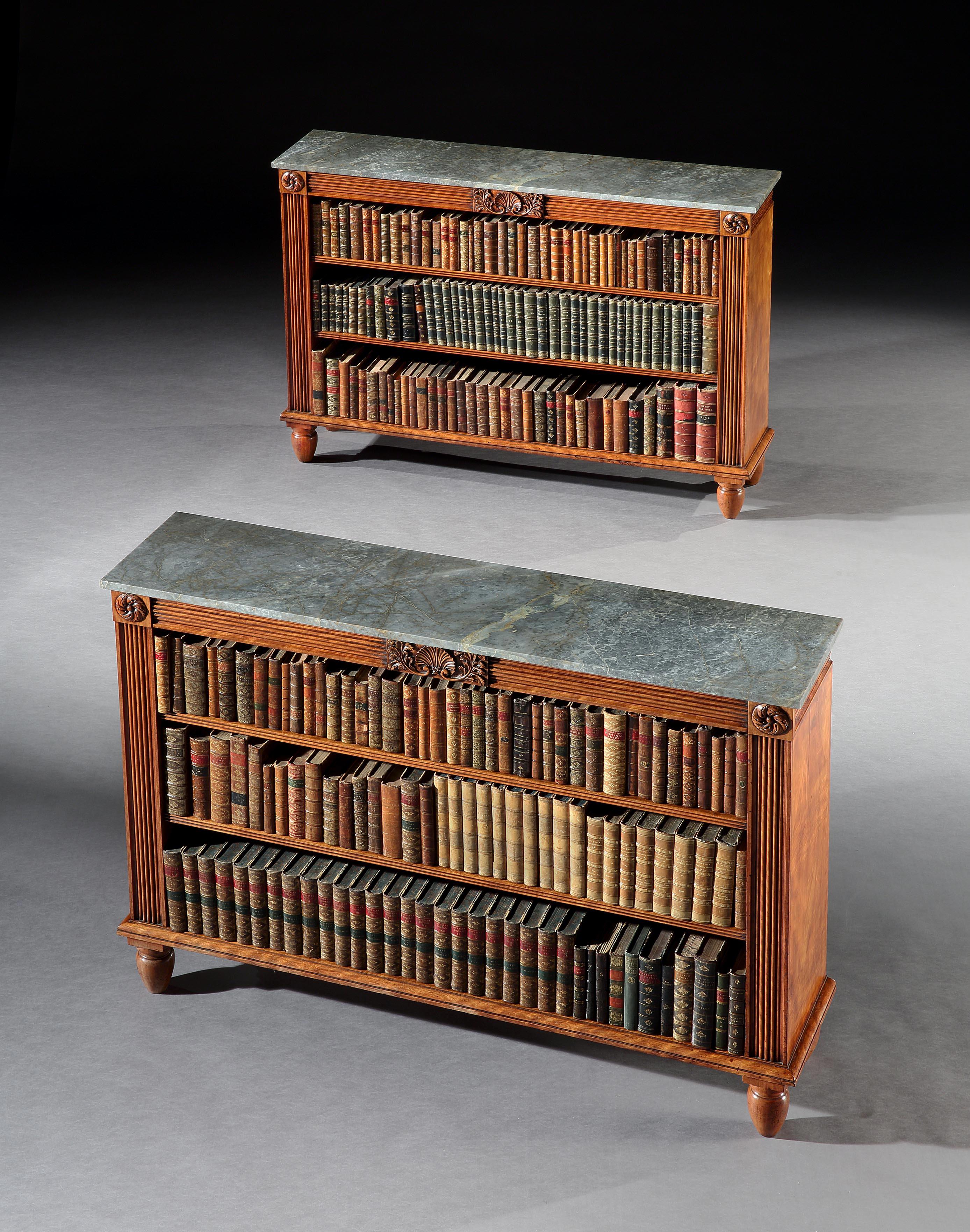 A pair of George III Regency open bookshelves in satinwood with monte carlo grey marble tops above a reeded frieze centred by a well carved shell and flaked by roundels above reeded pilasters and standing on turned feet.

  