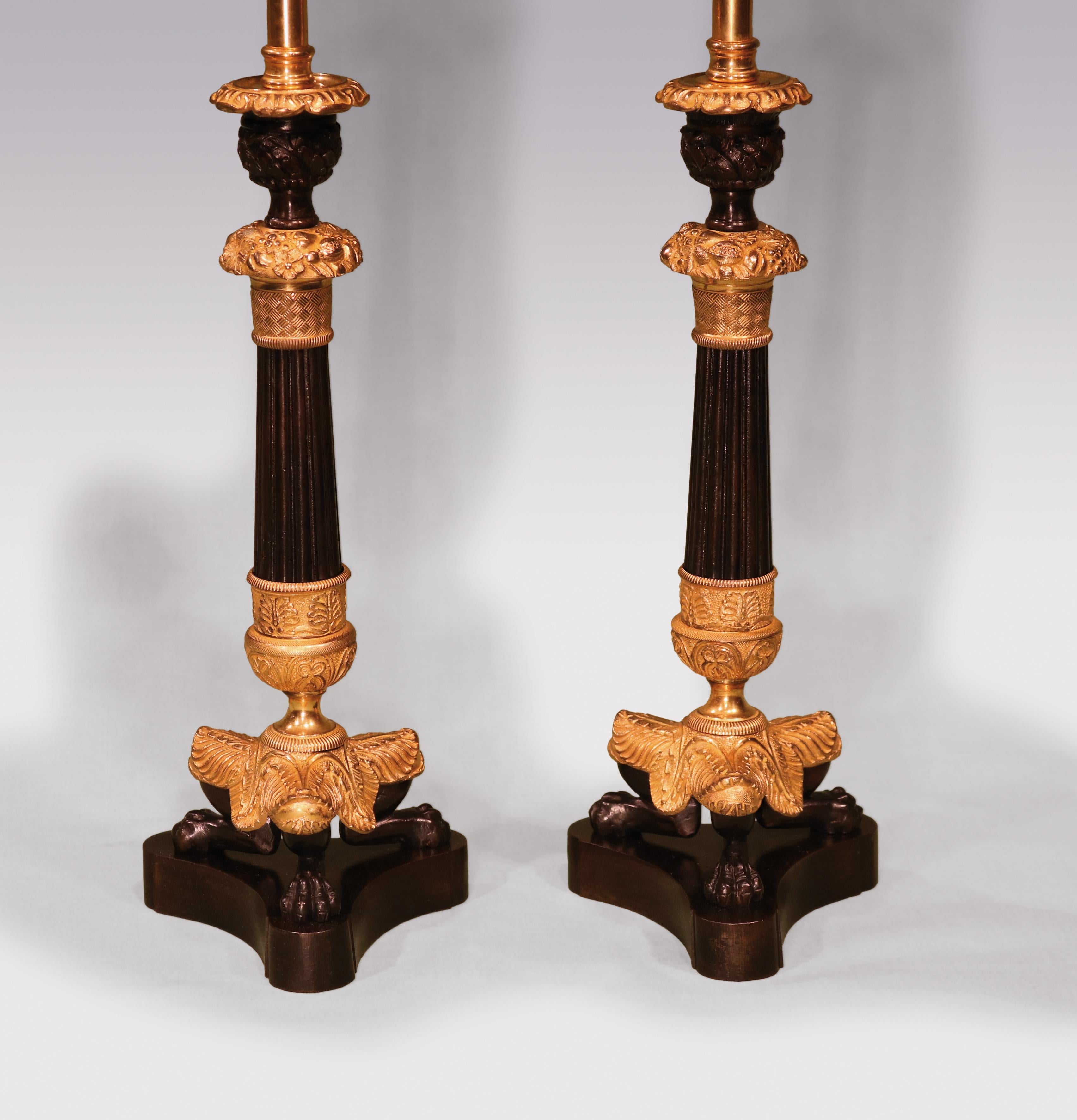 A pair of early 19th century bronze and ormolu candlesticks having urn shaped nozzles above engine turned and acanthus collared reeded tapering stems raised on leaf scroll, lions paw feet ending on concave platform bases. (now converted to lamps)