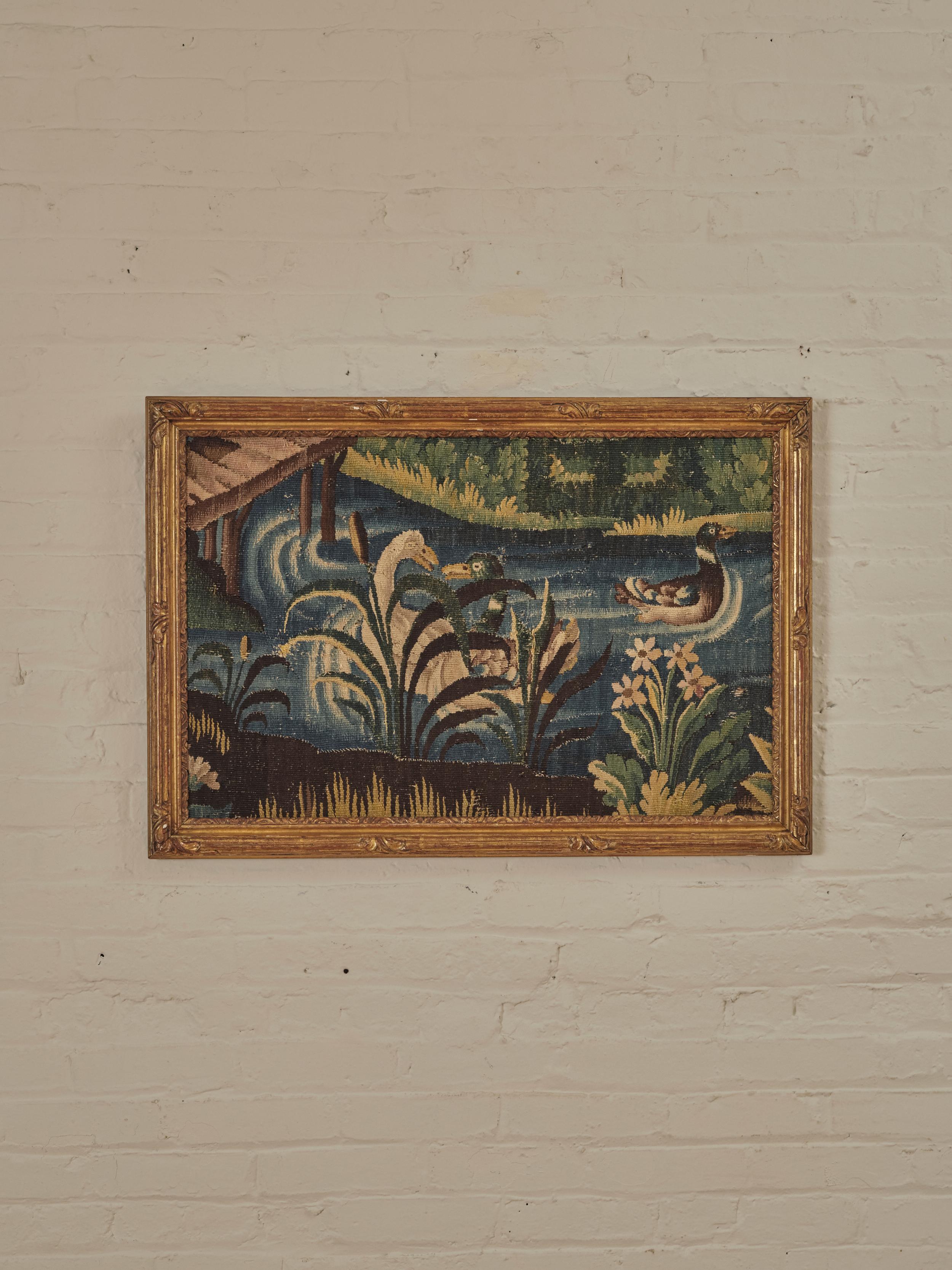A Pair of Riverside Landscape Framed Tapestries. Each fragment has been meticulously worked in multicolored wool and paints a riverside scene, complete with charming ducks and lush vegetation. The artworks are encased in carved and gilt wooden