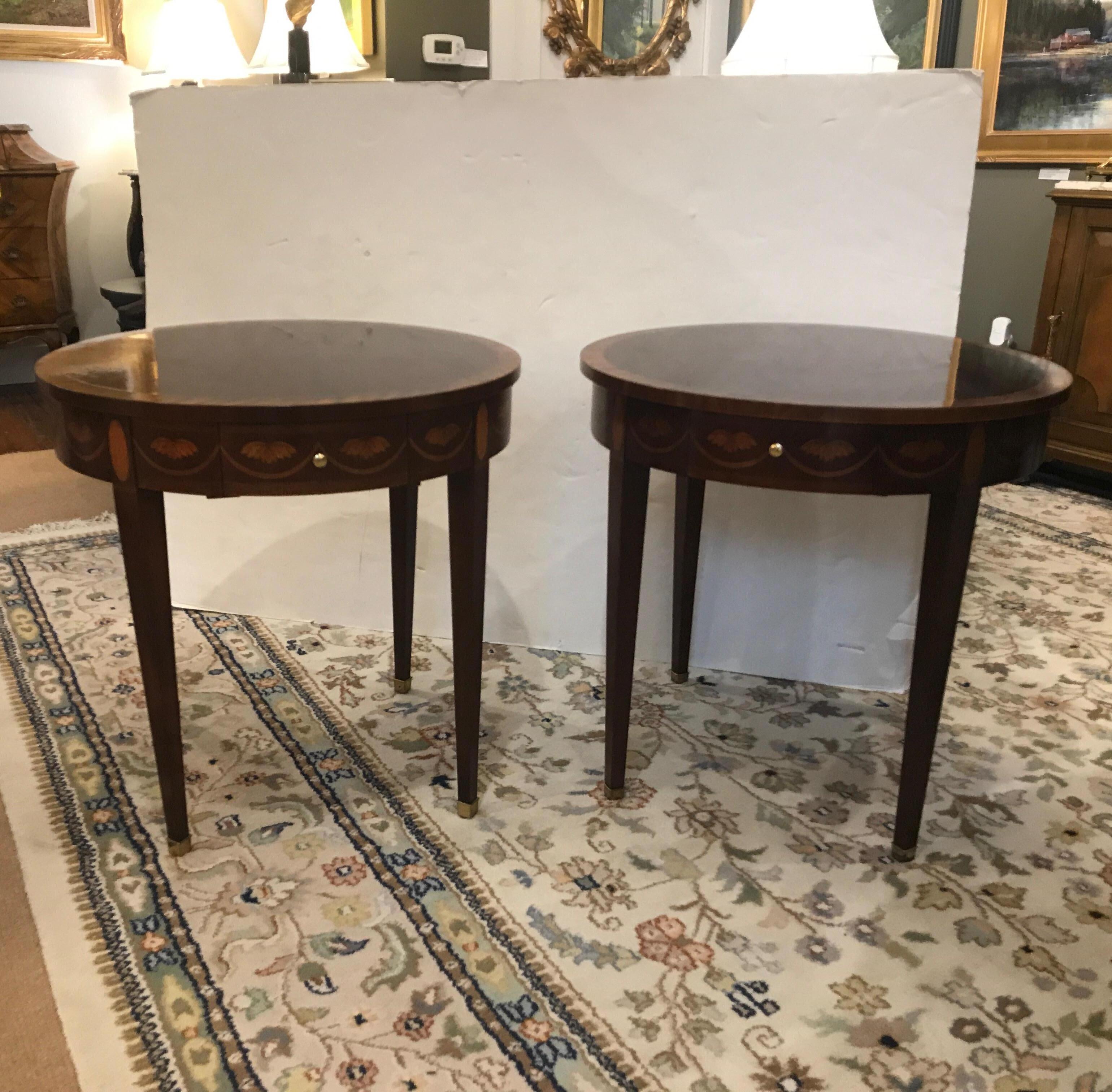 A pair of flame mahogany round side tables by Baker Furniture. The round tops with satinwood banding with side pullout / pull-out trays. The table with delicate inlay around the aprons and the top of the four tapering legs. Each one with a baker