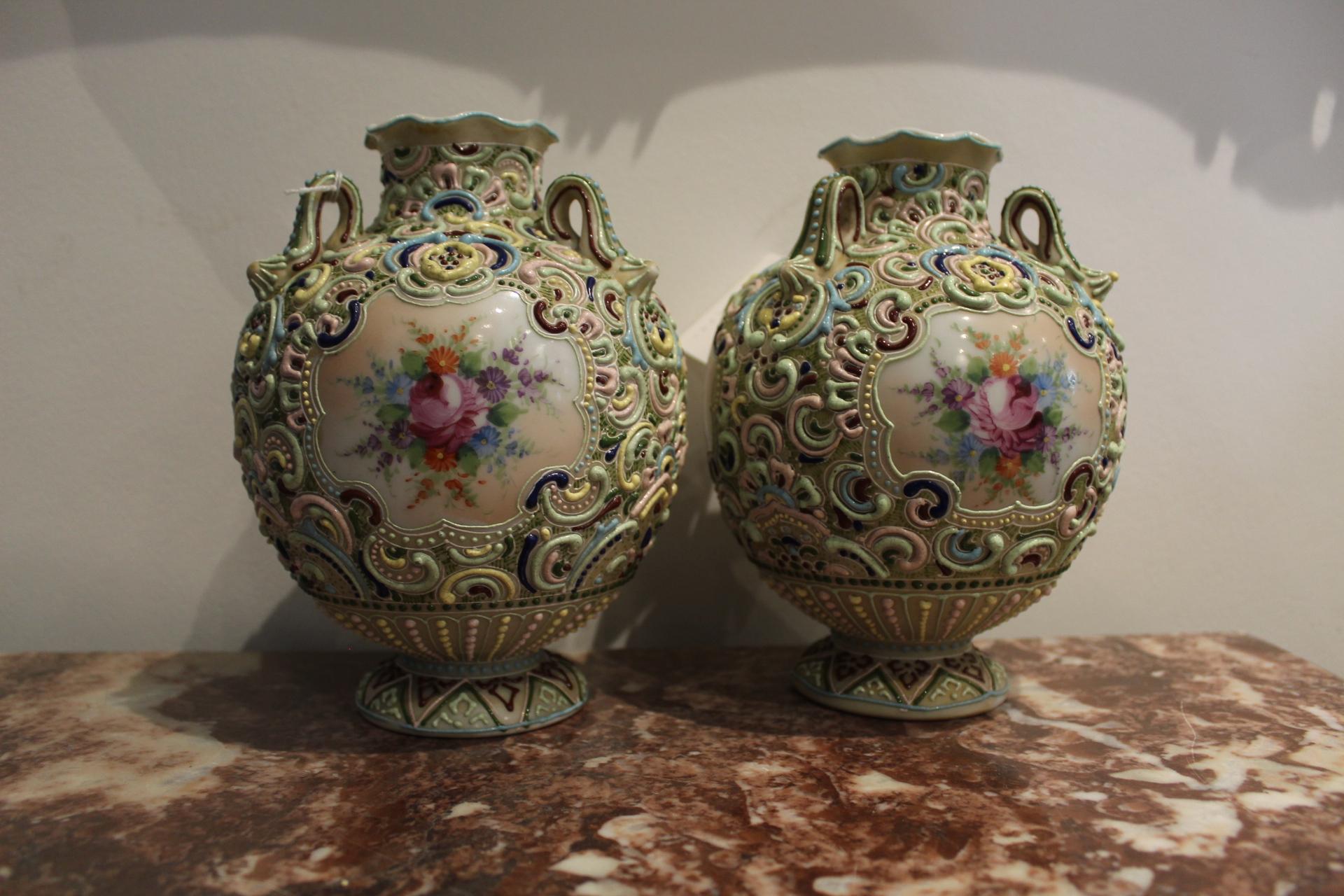 The pair feature a round foot upon which a globular body sits with a short gently ruffled neck, surrounded by three small decorative handles. Having a matte body, they are decorated in a thick colorful decoration using diluted clay or slip, called