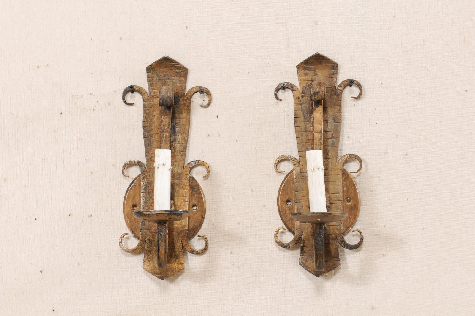 A pair of Spanish gold-tone iron sconces from the mid-20th century. This vintage pair of sconces from Spain each feature a stylized fleur-de-lis body with a single arm extending out into a downward swag, supporting a ruffled iron bobèche and painted
