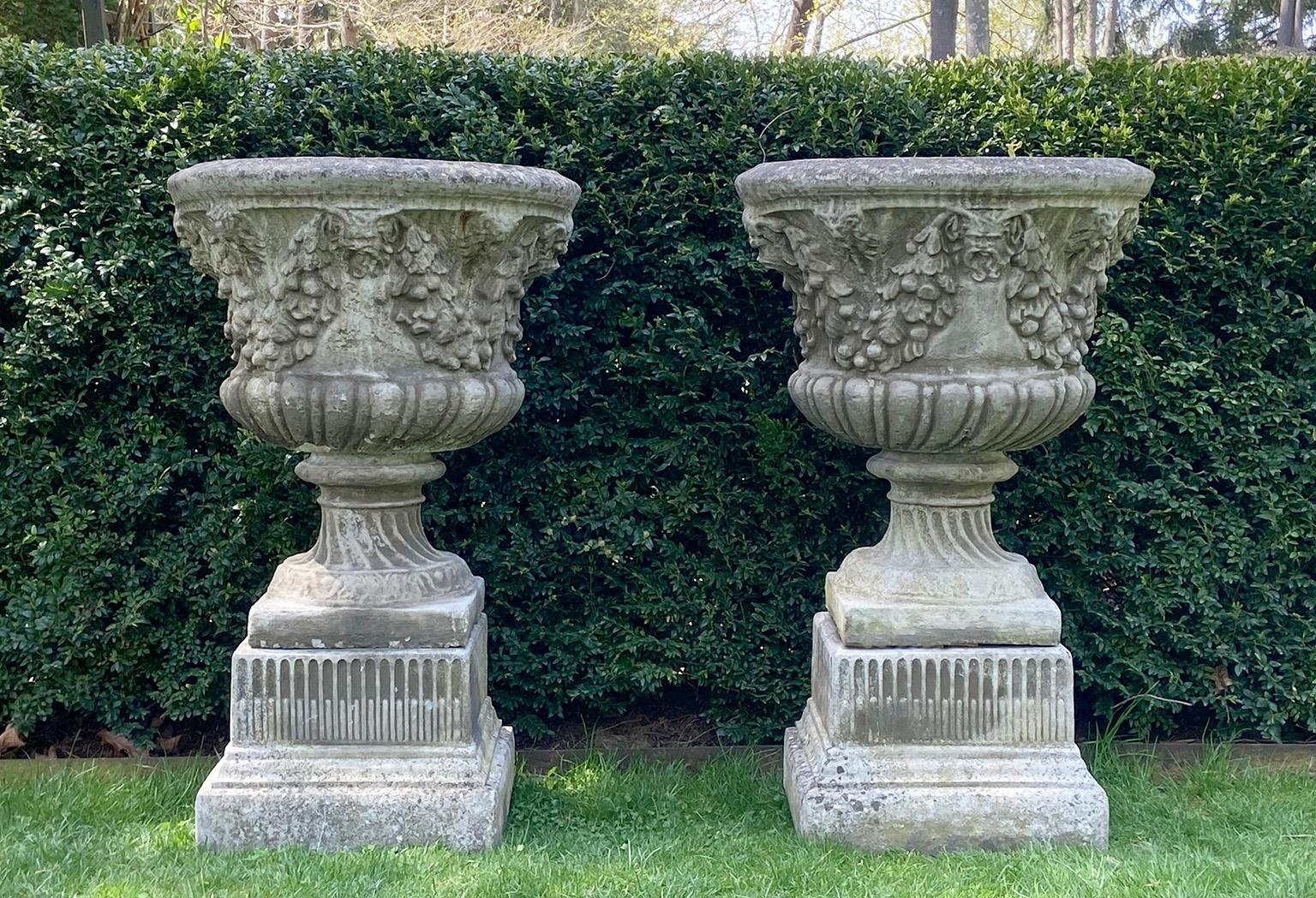 A pair of composition stone urns, each with partially-lobed body, the upper portion of the body ornamented with fruited swags and grotesque masks, on wrythen, fluted, circular socles and integral square bases, each atop square partially-fluted