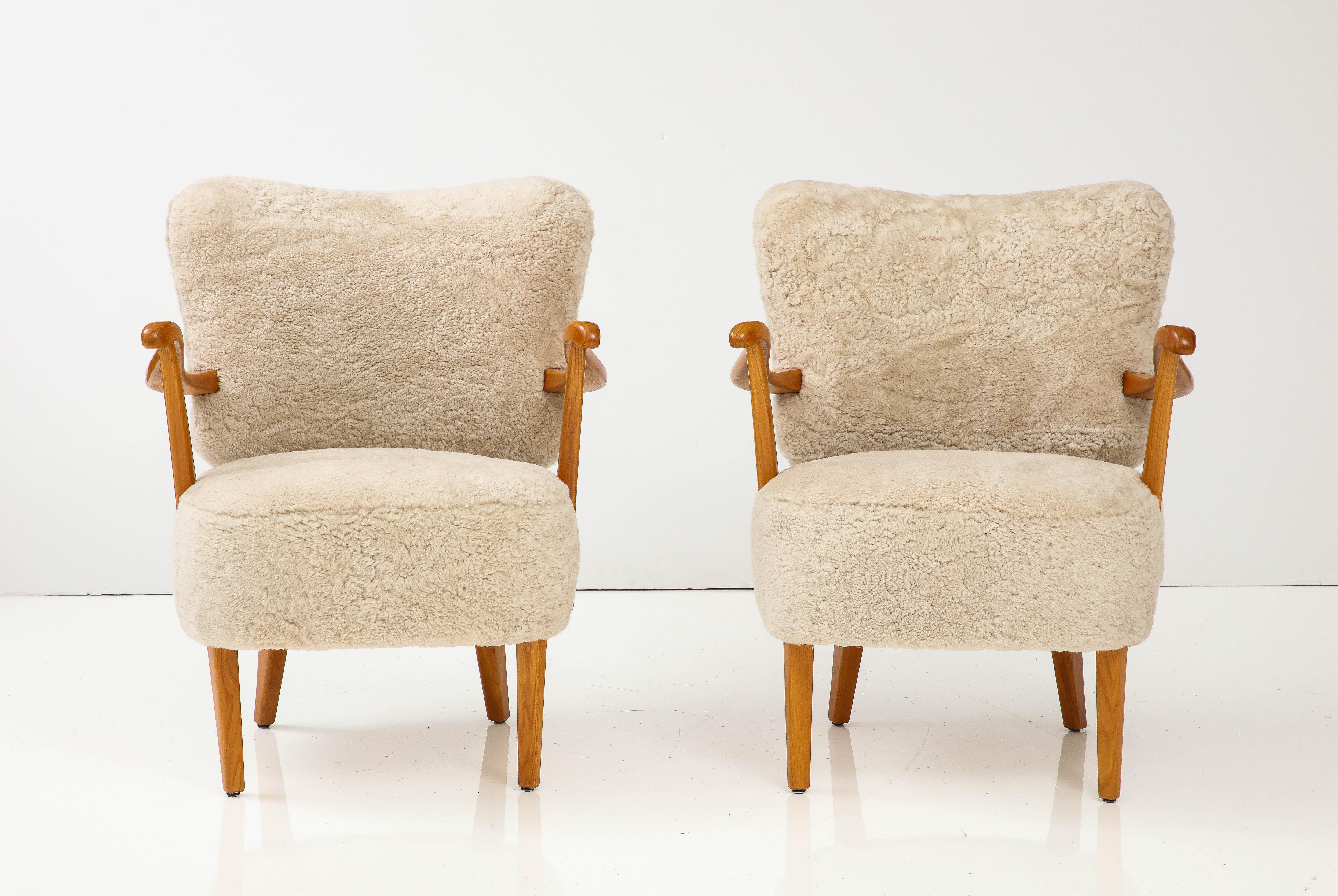 A pair of Swedish Modern elmwood open armchairs, Circa 1940s, with the curved sheepskin backs, scolded armrest above upholstered seats raised on tapered legs.
