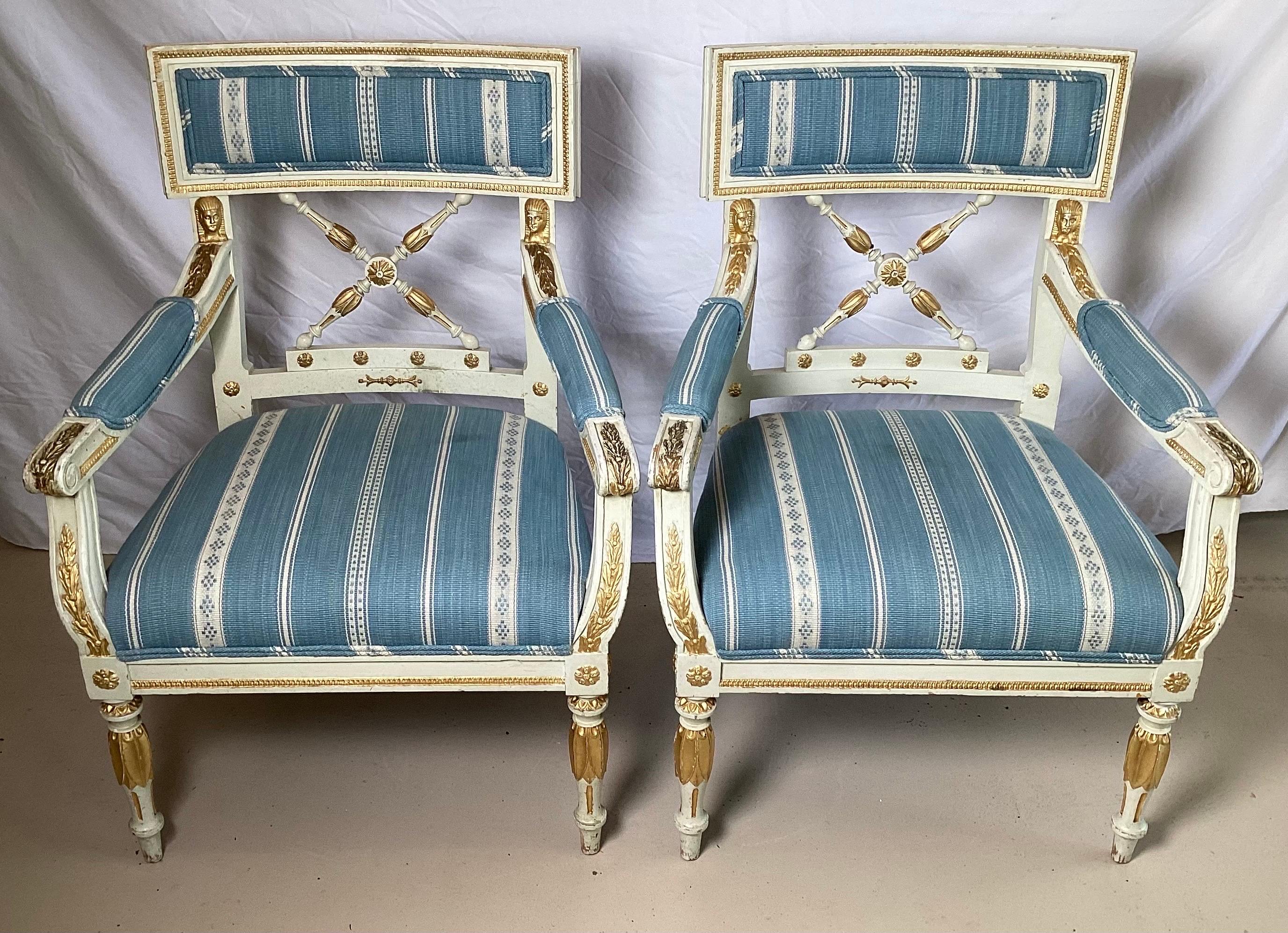 A pair of white and gilt paint decorated open arm chairs. The Napoleonic themes frames with new blue and white striped fabric.