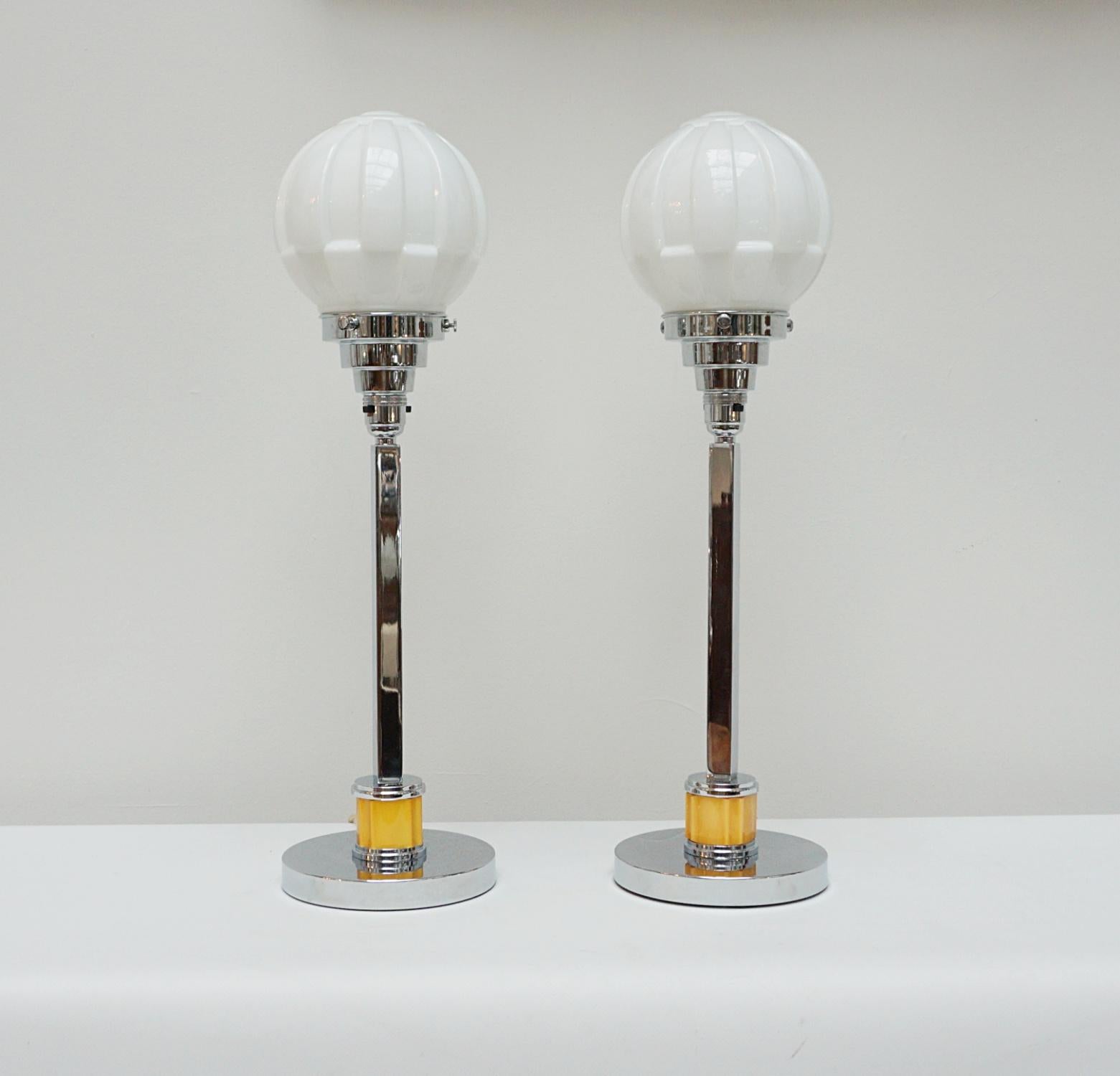A pair of tall Art Deco table lamps. Chromed metal squared stems with a section of yellow bakelite. Set over stepped circular base. White glass globe shades. 

Dimensions: H 55cm W 16cm 

Origin: English

Item Number: J289

All of our lighting is