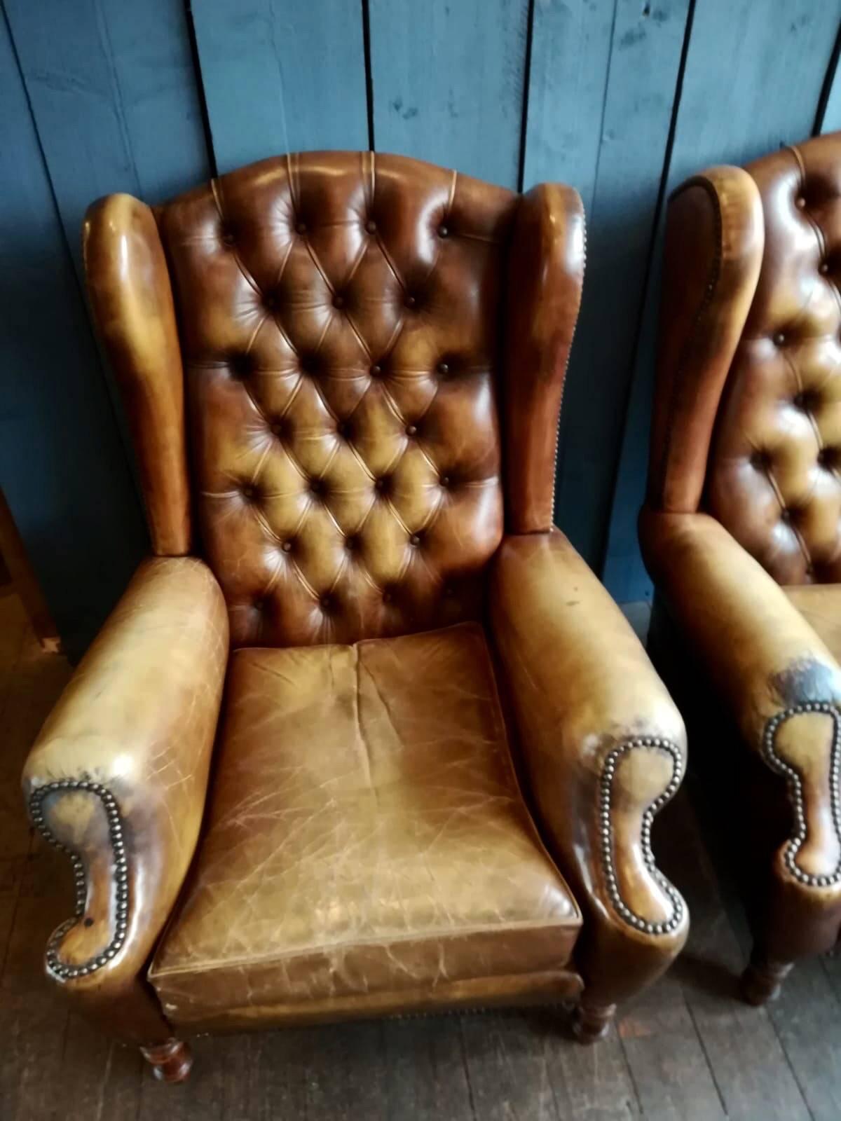 A nicely worn pair of vintage tan leather wingback armchairs. Having a button back and a nice stud detail. Standing on turned beech legs.