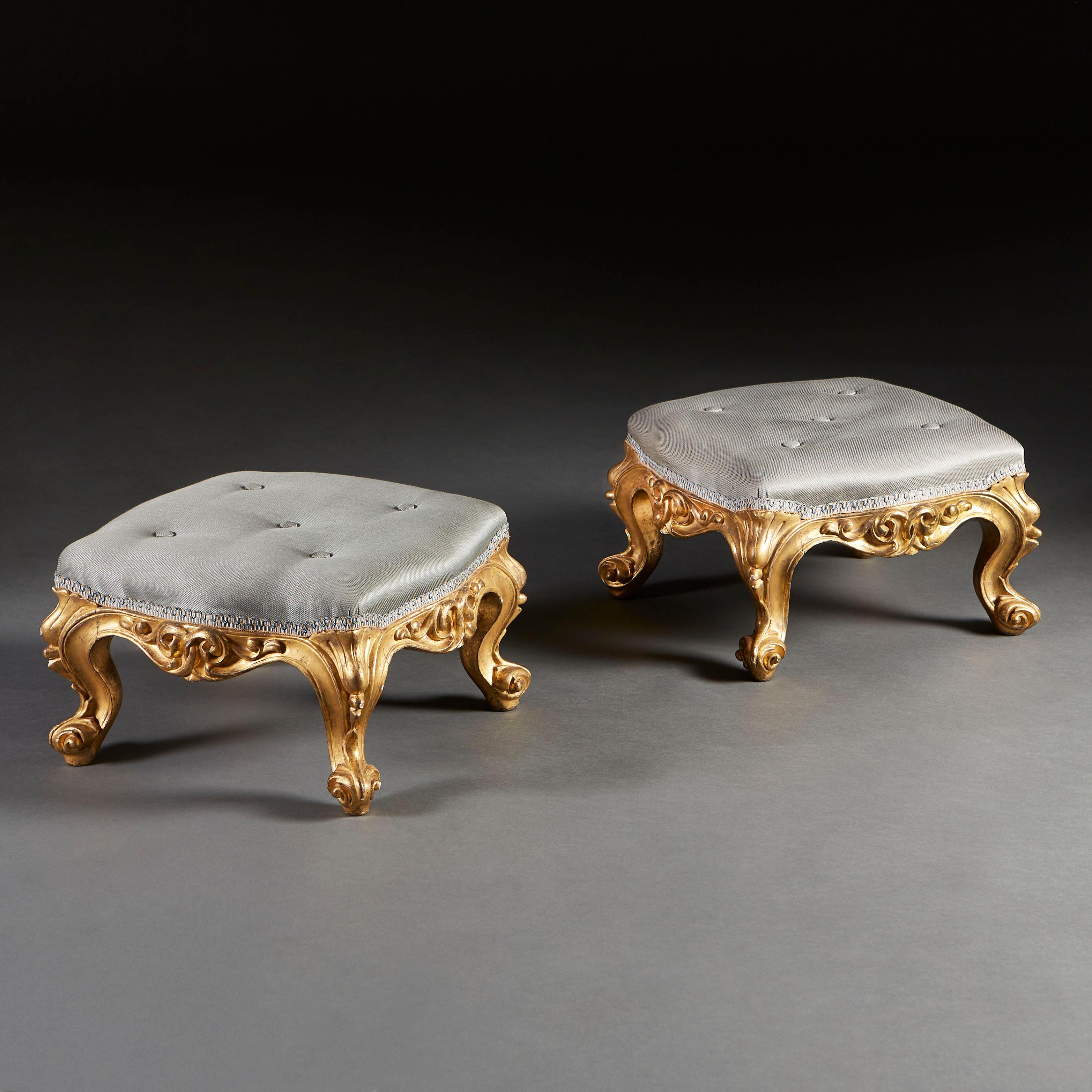 A fine pair of William IV giltwood footstools, with scrolling carving and cabriole legs with scroll feet, upholstered in blue fabric with float buttons.