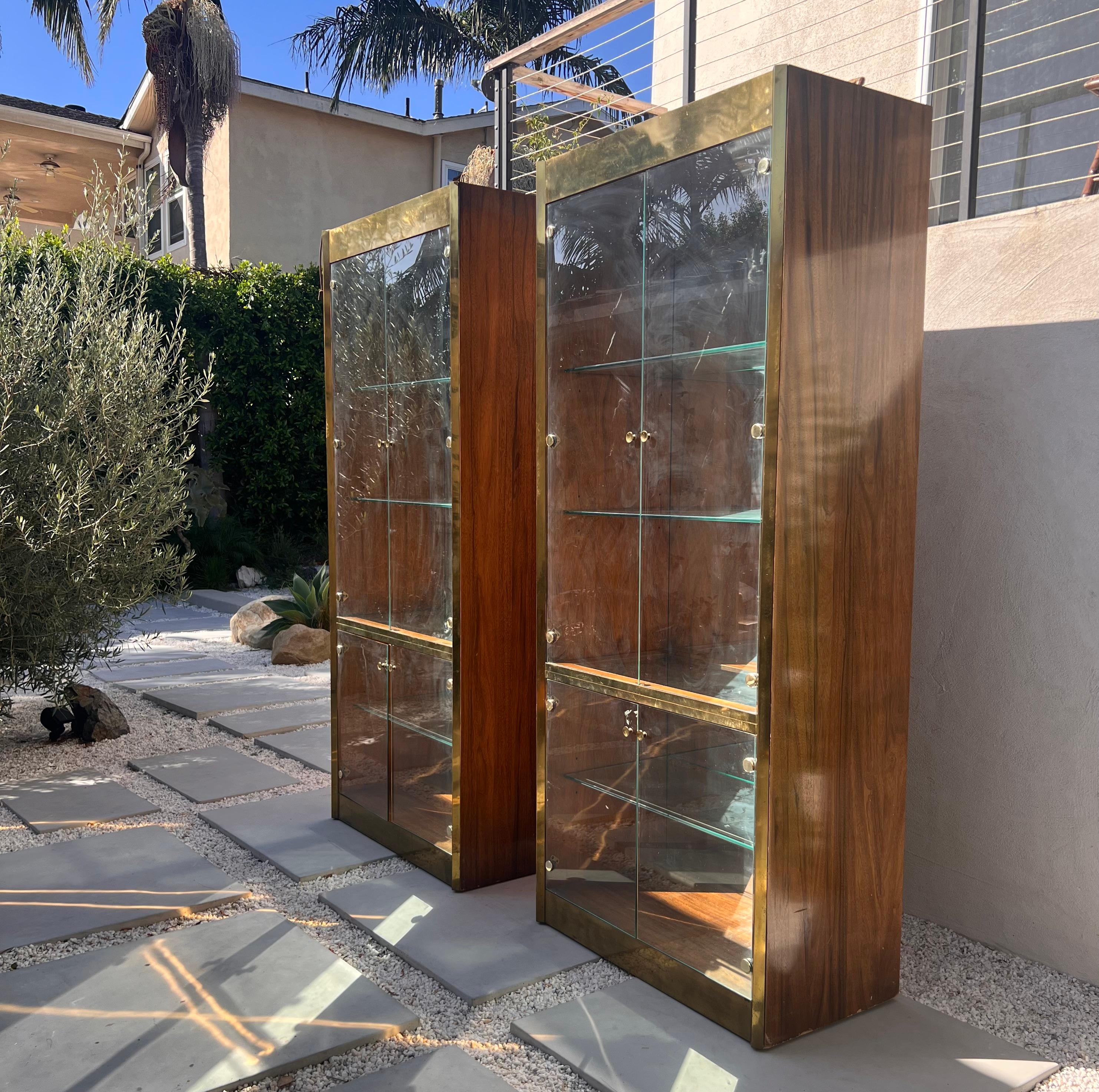 A pair of mid century modern monumental étagères / display case cabinets by Mastercraft, circa early 1970s. Wood with brass detailing and hardware and featuring mirrored backs and glass shelves, as well as lights on the interior of the ceiling of