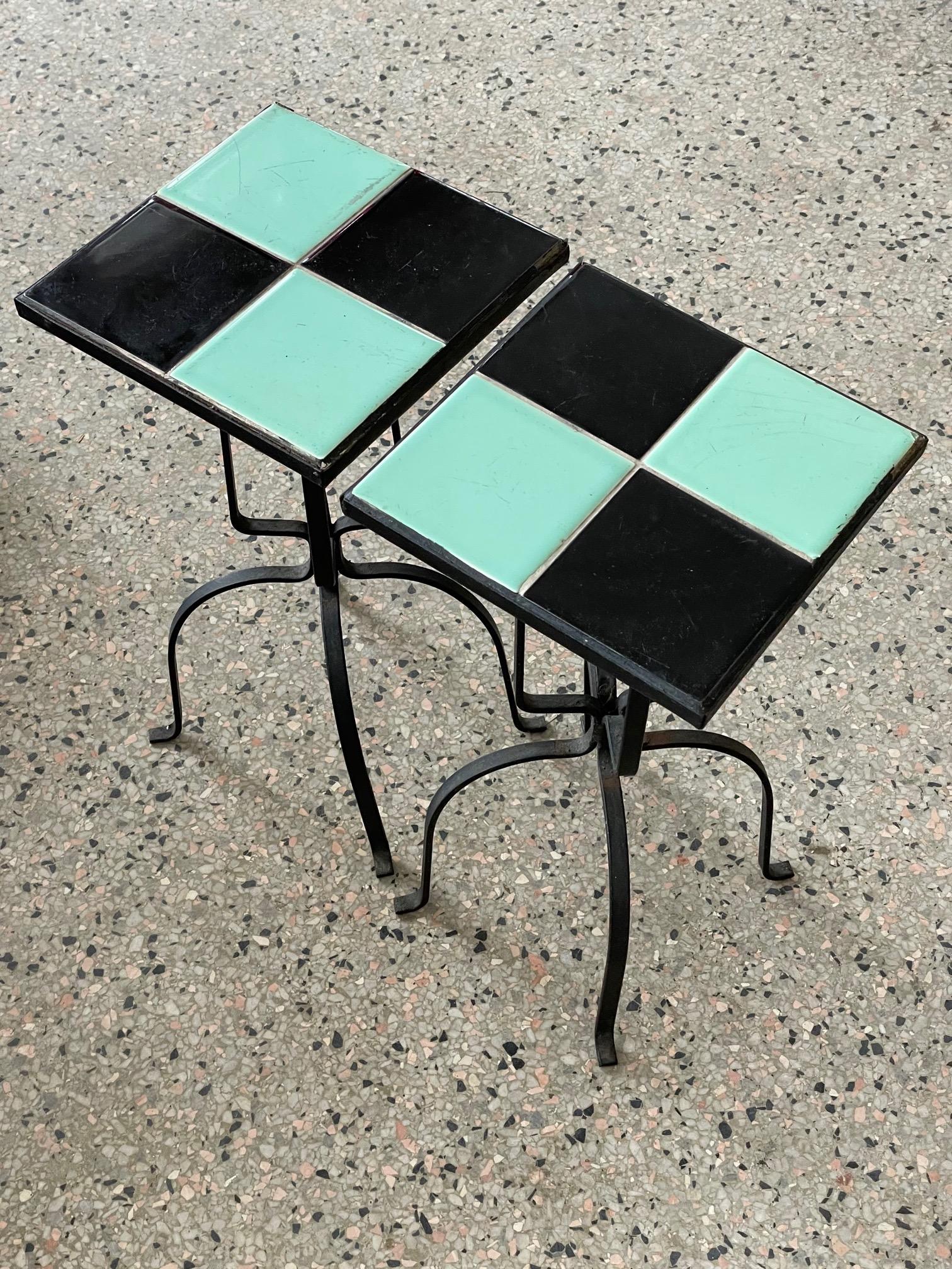 A pair of wrought iron and tile top tables. The tiles are black and blue/green. The tables are 18
