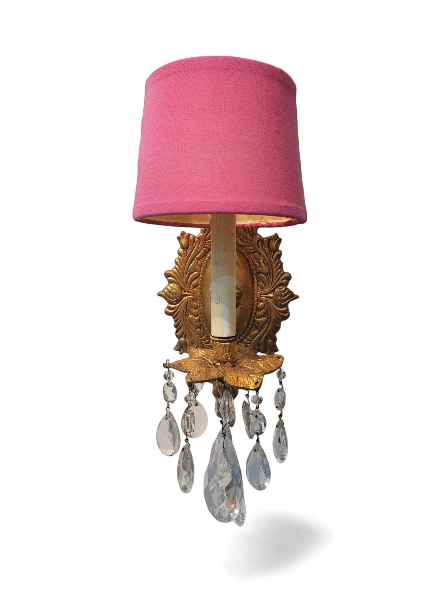 A pair of elegant, electrified wall sconces featuring vibrant pink shades and ornate gilded metal backplates in a floral sunburst motif. Each sconce is adorned with cascading clear crystal prisms. 
Embodying the playful glamour of Palm Beach Regency