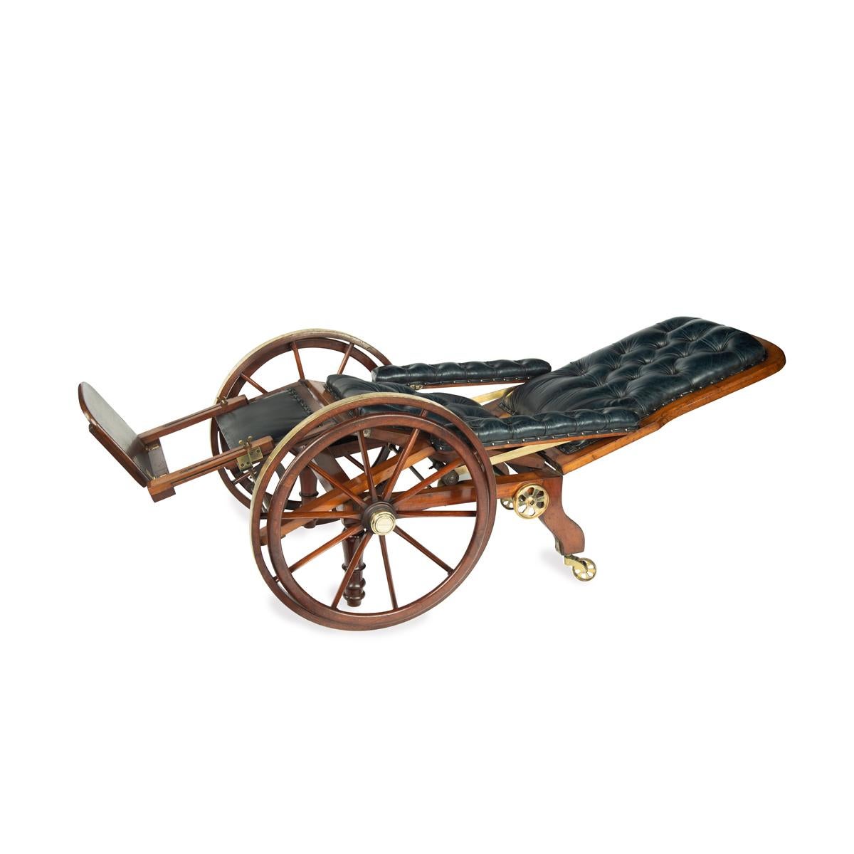 This mahogany wheel chair has a padded back, arms and seat, reupholstered in deep buttoned blue leather.  The brass-bound wheels have a secondary outer wheel, and a pair of smaller brass wheels which enable the chair to be reclined almost to the