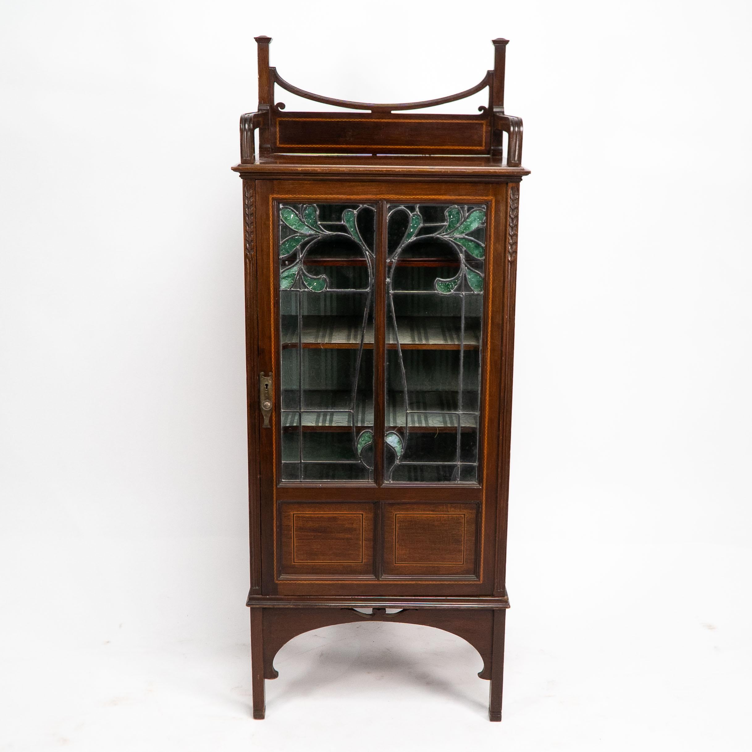 A petite Arts and Crafts mahogany display cabinet in the Anglo-Japanese style with chequer string inlays and carved fern details to the upper sides with stained and coloured leaded floral glass work to the door.
 
 