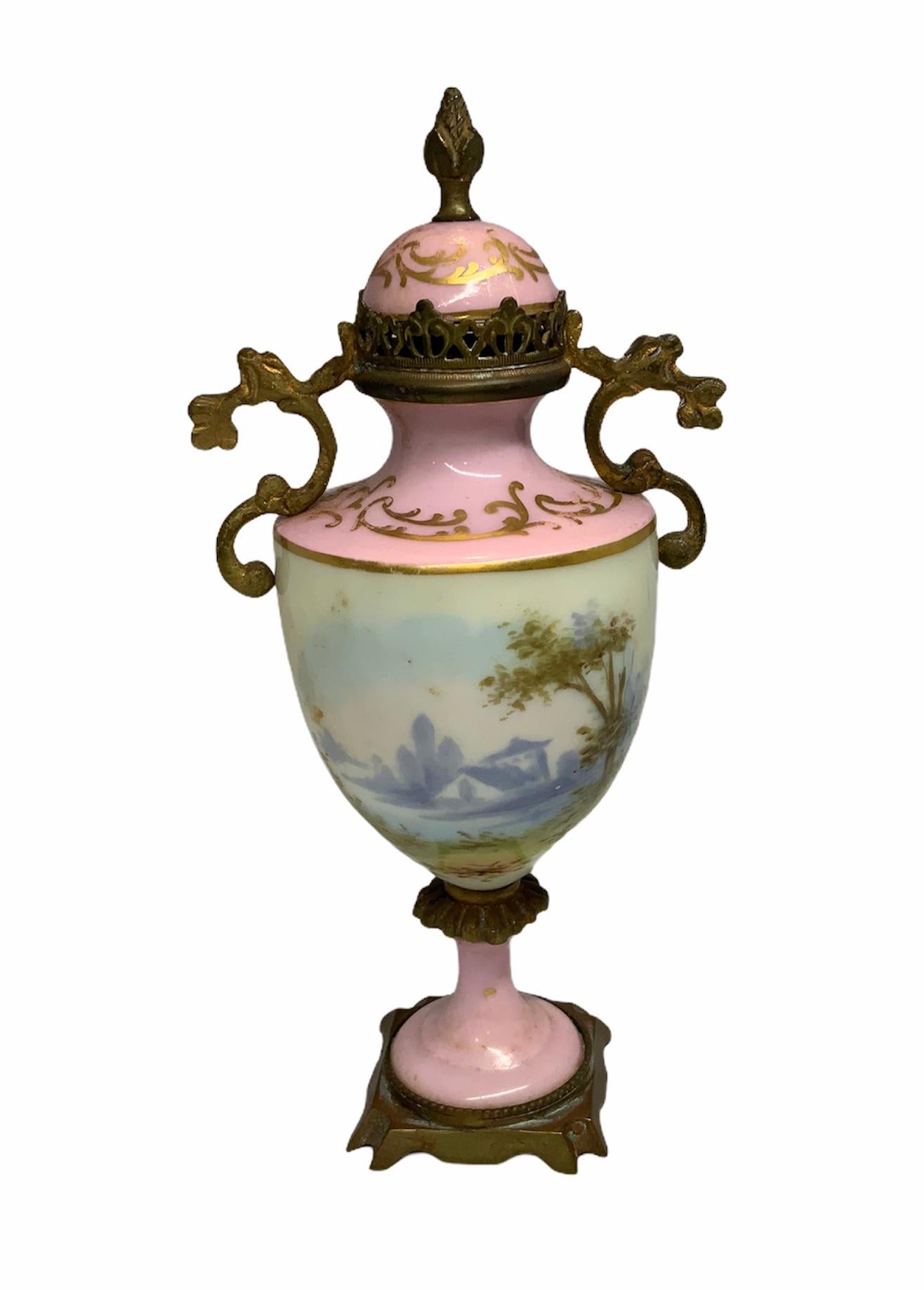 This is a very old gilt and pale pink background urn depicting a continuous hand-painted scene of a lady sitting in a pastoral landscape holding her fan and observing nature. In the back there is a scene of a lake in front of a country house. The
