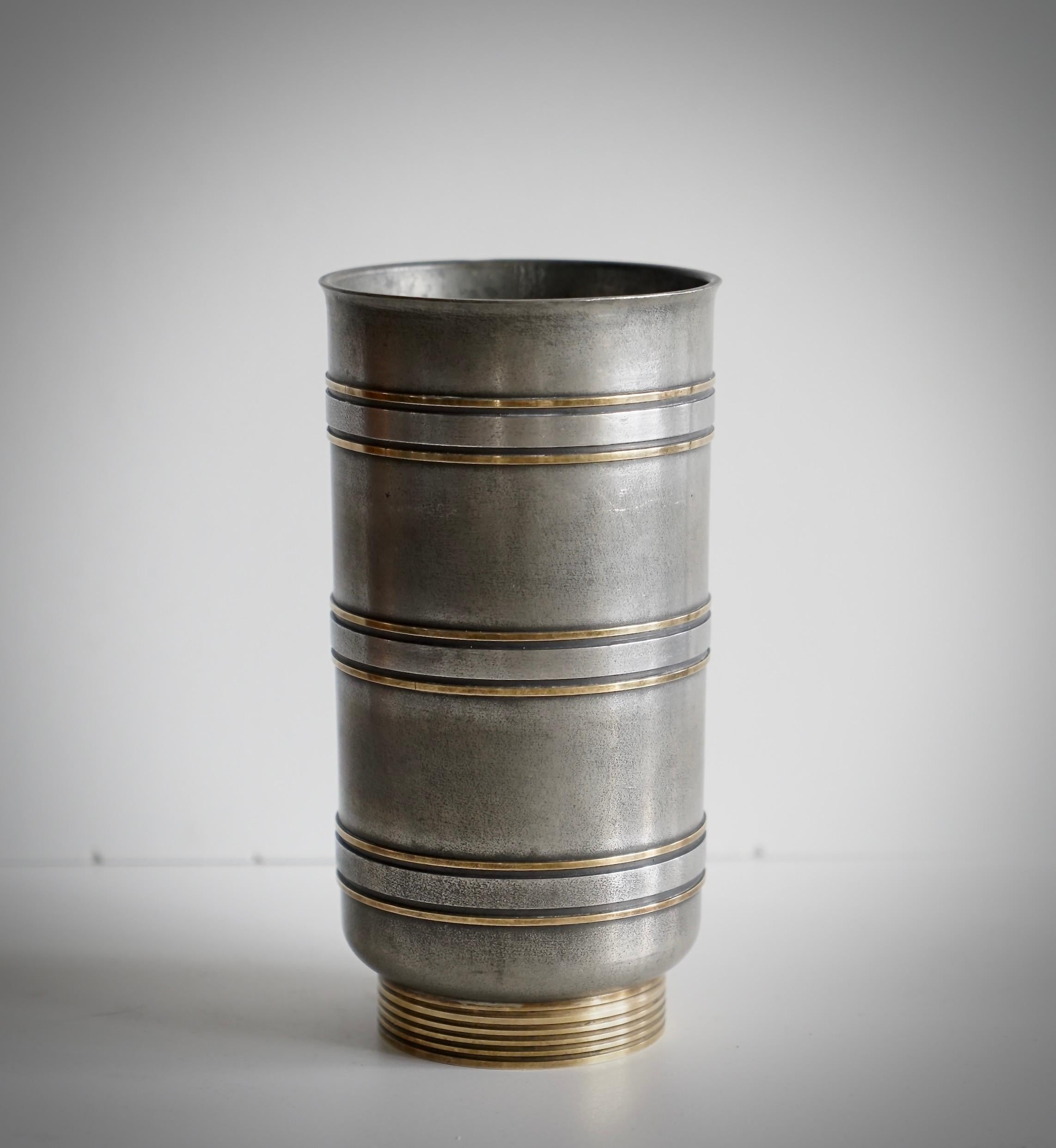 Vaso a forma di cilindro in peltro  con tacco scanalato decorato e fascia in ottone. Disegnato da Edvin Ollers (1888-1959). Timbro OLLERS, S&O Schreuder & Olsson, CAST TIN e timbro dell'anno R8 per il 1943.
Piccola ammaccatura sul bordo superiore.