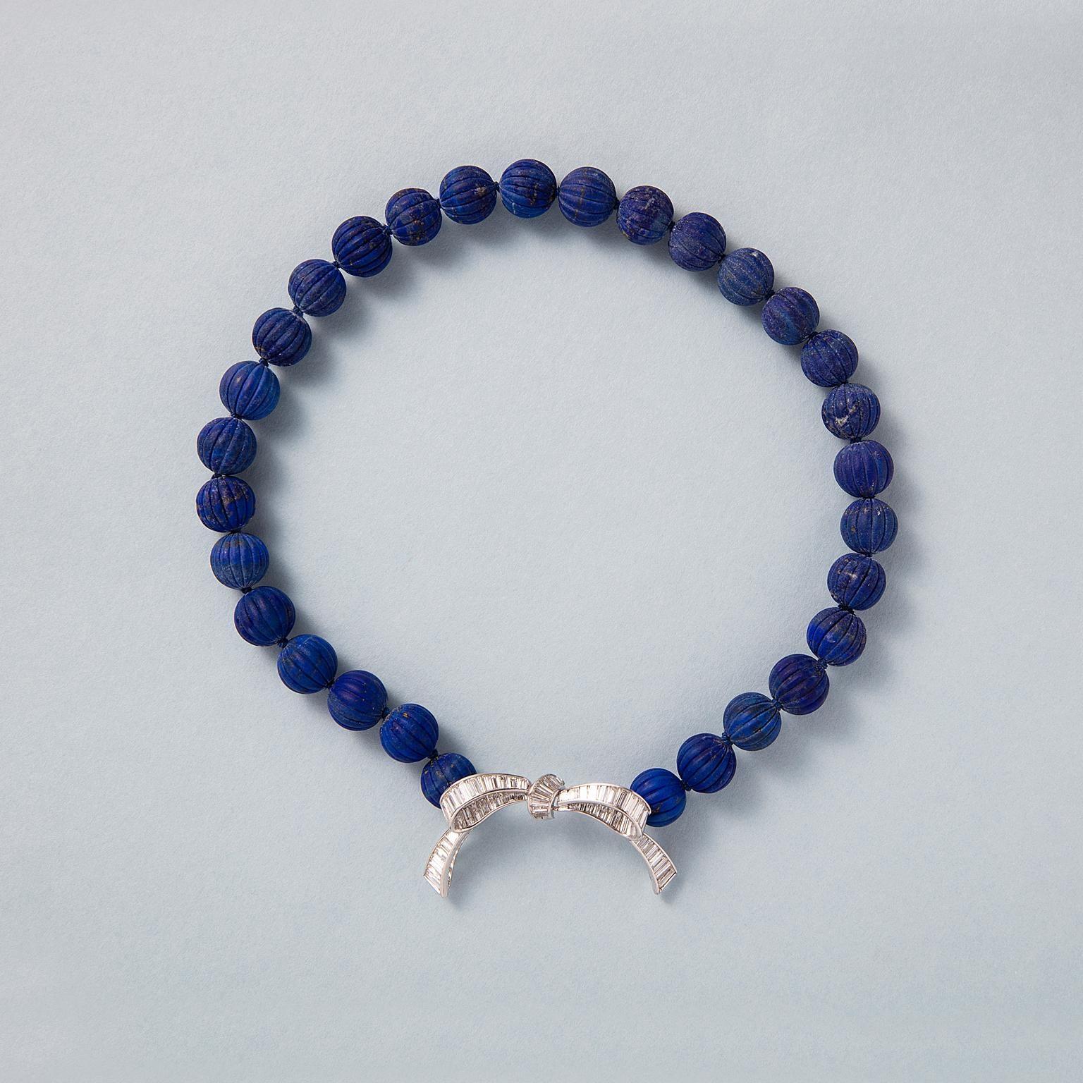 Un elegante fiocco in platino e diamanti si appoggia su una collana a girocollo con perline di lapislazzuli taglio melone. Il fiocco è incastonato con circa 8 carati di diamanti taglio calibré incastonati a canale e taglio baguette, americano, circa