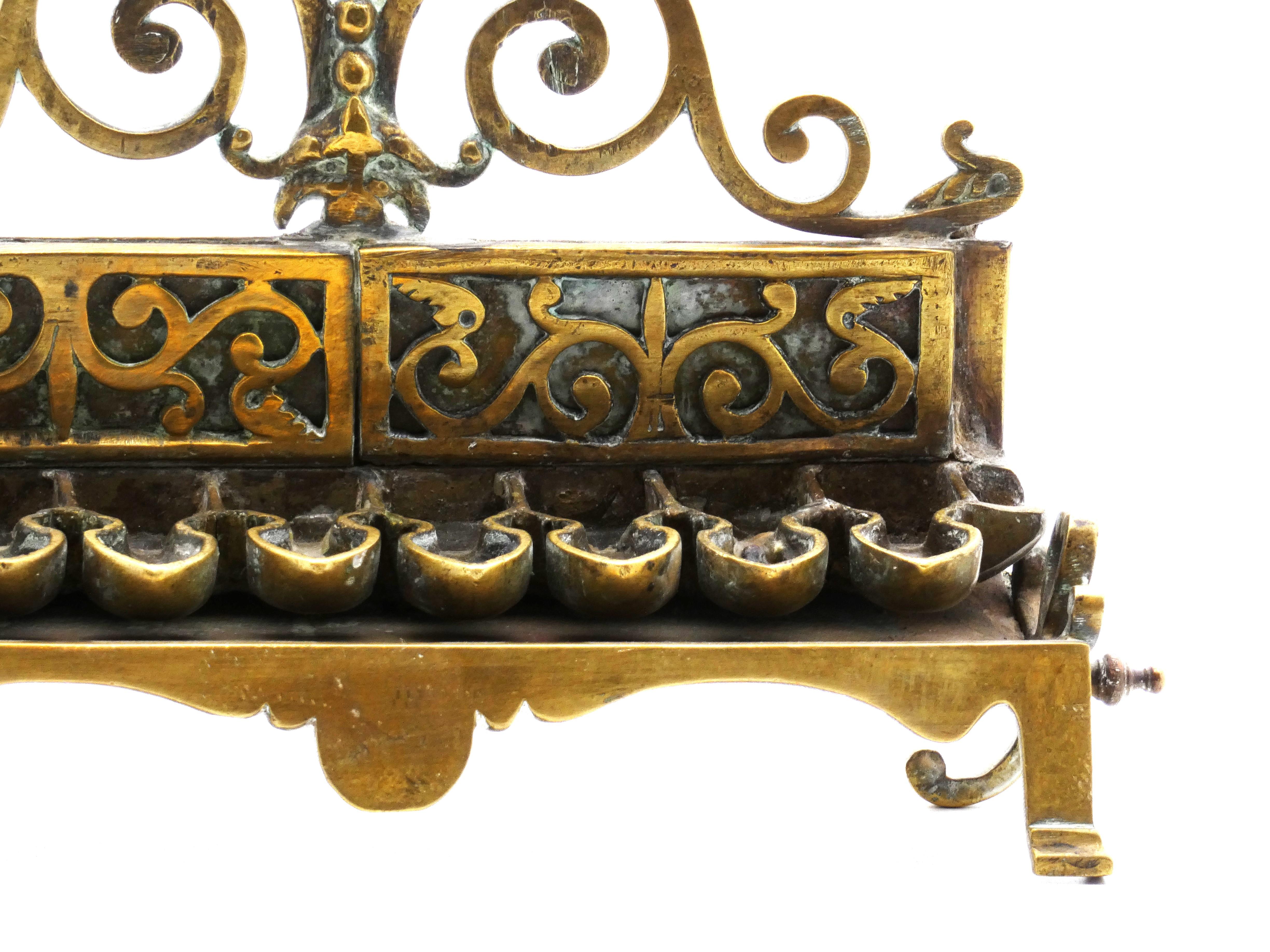 Hanukkah Lamp is interestingly made with many different classical shapes and parts. 

At top, a pierced fleur de-lis surmounted on scrolling rails is mounted atop a similarly pierced backplate attached to the main frame. 

Each square lamp