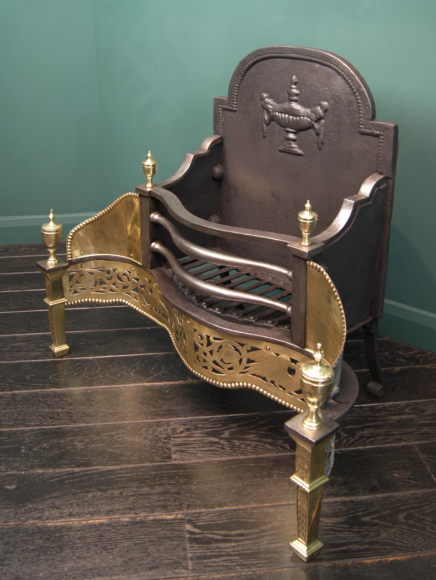 A polished brass decorative dog grate of serpentine form, the shaped wrought fire bars over pierced openwork fret set between tapering uprights. The decorative fireback with central urn detail. Extensive engraving to fret, wings and finials and