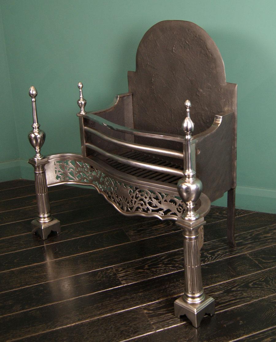 A polished steel and cast-iron fire grate of Chippendale form. The railed serpentine front above a pierced and engraved frieze with dragon and floral detail amidst foliate scrolls, flanked by fluted column uprights with elongated urn finials on