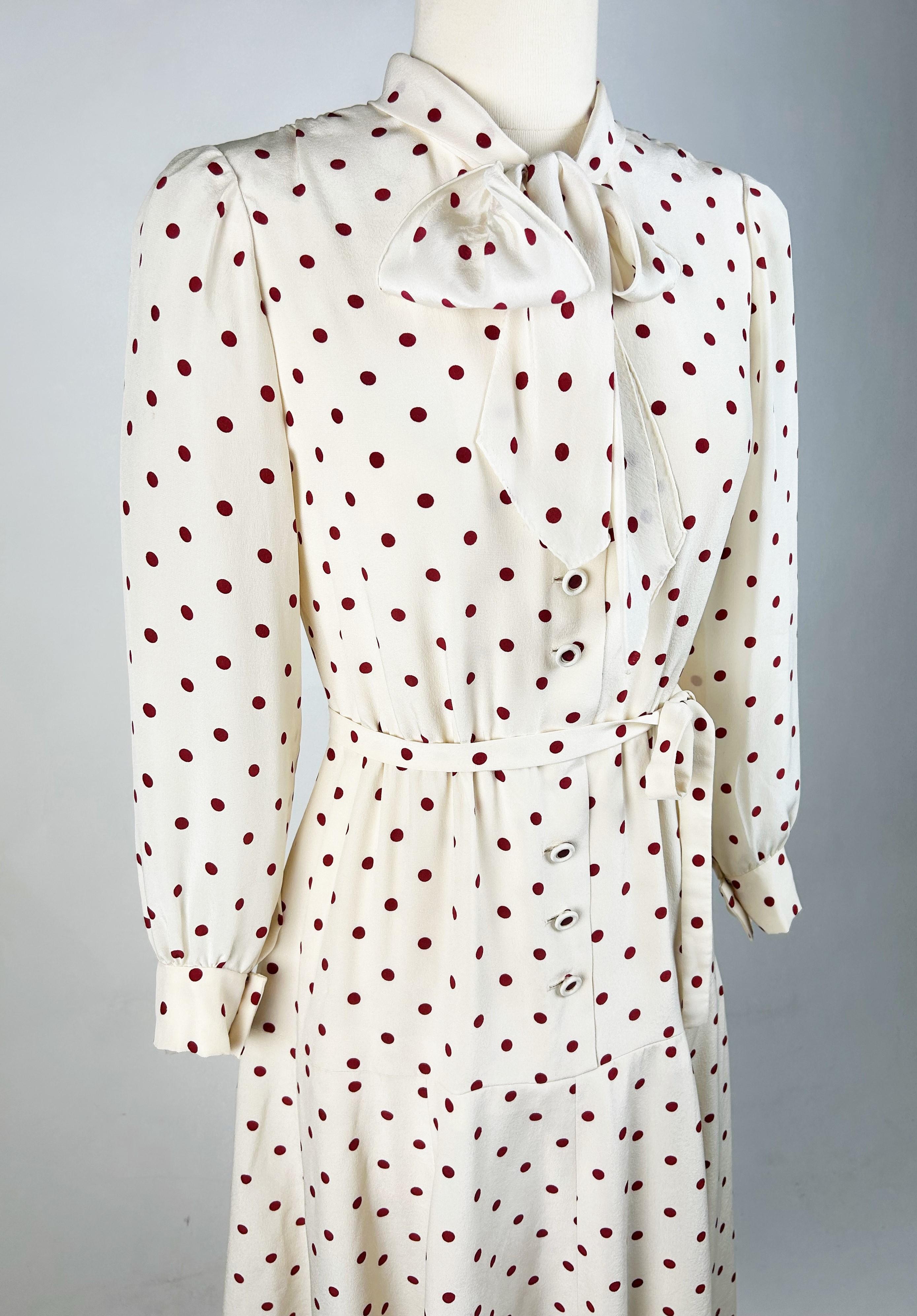 Circa 1970-1980
France
Chanel Haute Couture cocktail dress numbered 59644 dating from the 1970s. Short dress, long sleeves with small puffs, in ivory silk crepe printed with deep red polka dots. Ascot collar with ganser front and nine matching