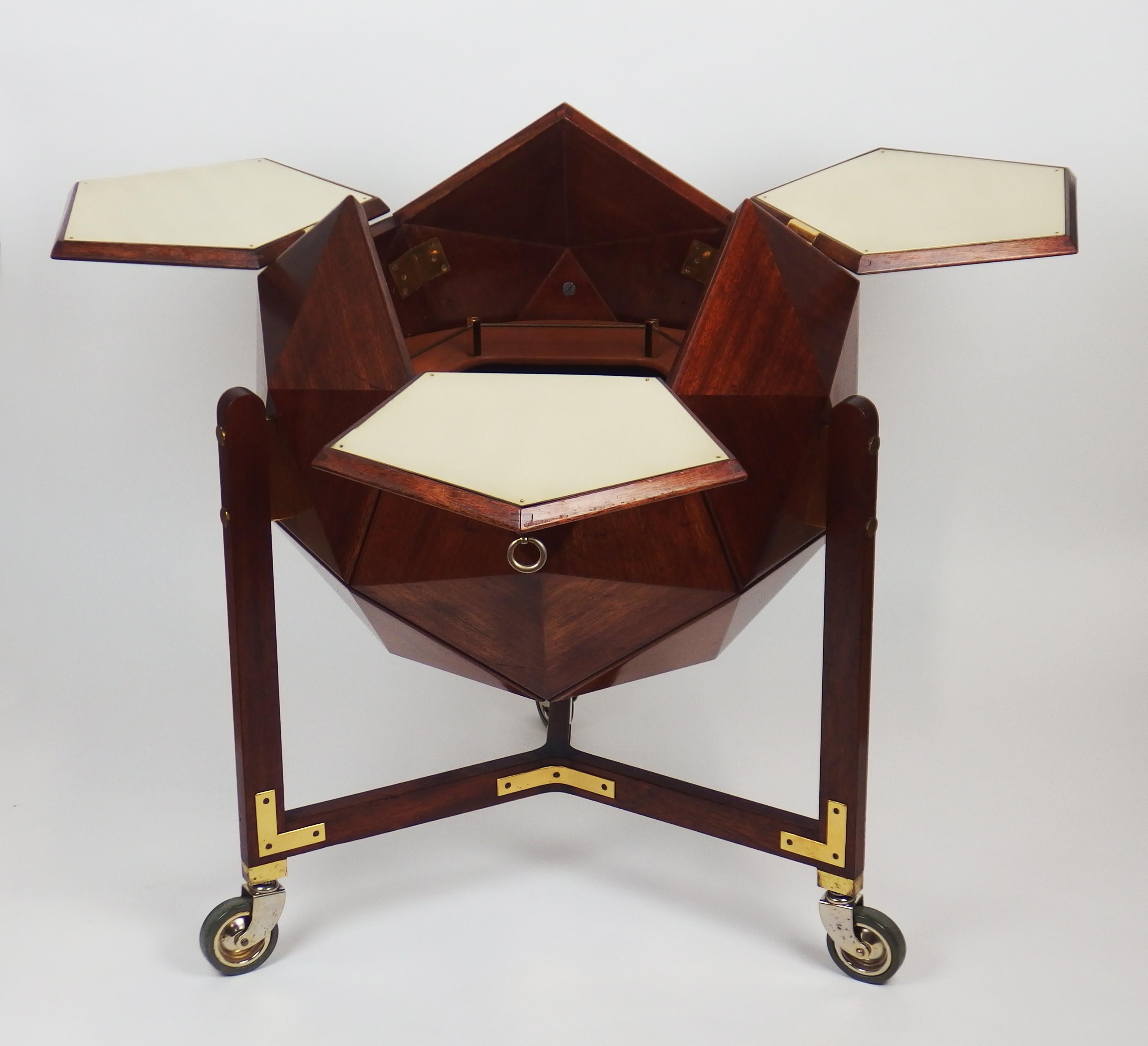 A polyhedron shaped mahogany bar on castors with plated brass and brass details. Three drop down doors opening to a place with a circular brown moleskine shelf for stocking glasses and bottles.