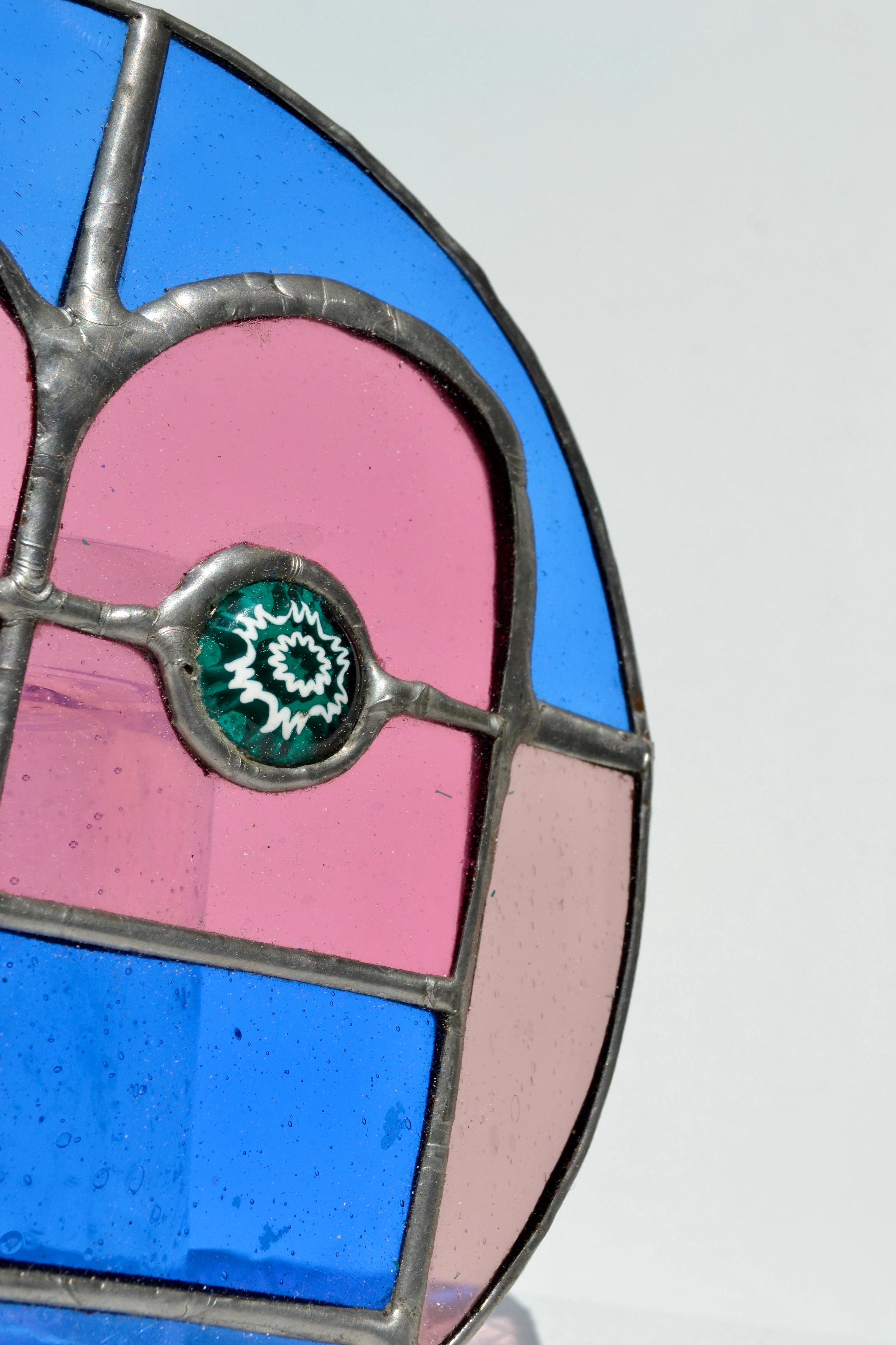 Excepcional panel pequeño de vidrieras de los años 80, hecho a mano con vívidas piezas de vidrio rosa y azul, acentuadas por intrincados detalles millefiori verdes.

Esta pieza redonda única captura el espíritu alegre y vanguardista del diseño