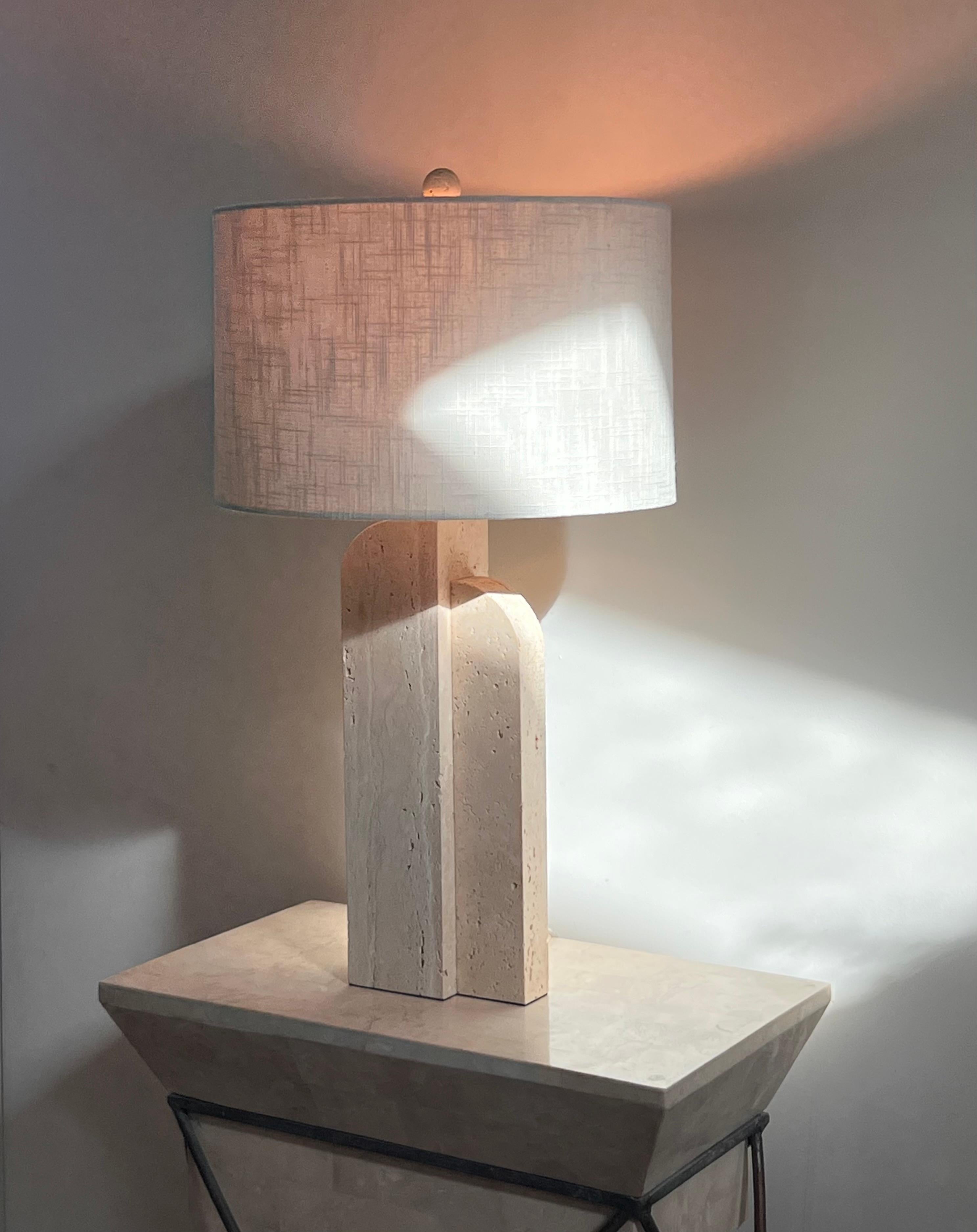 A postmodern travertine table lamp with canvas drum shade, late 20th century. Comprised of two asymmetrical arched blocks of travertine, this piece is at once blunt and whimsical, marrying the hardness of material with imaginative form. Having been