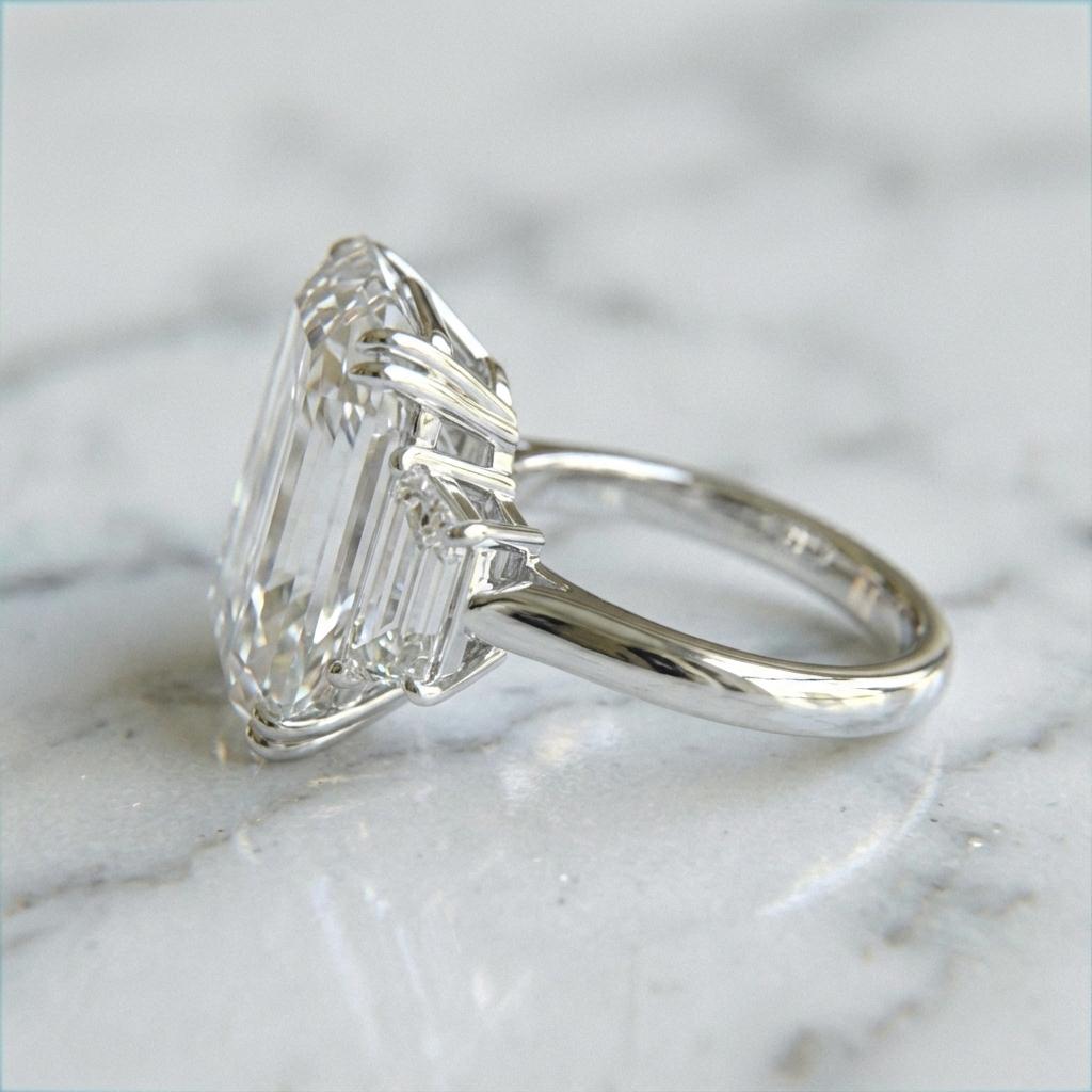 Crafted in shimmering platinum and framed by two tender trapezoid diamonds, this 9.13 carat Emerald Cut becomes more than a jewel—it becomes a quiet promise. With every long, graceful facet, it reflects a love that is steady, luminous, and