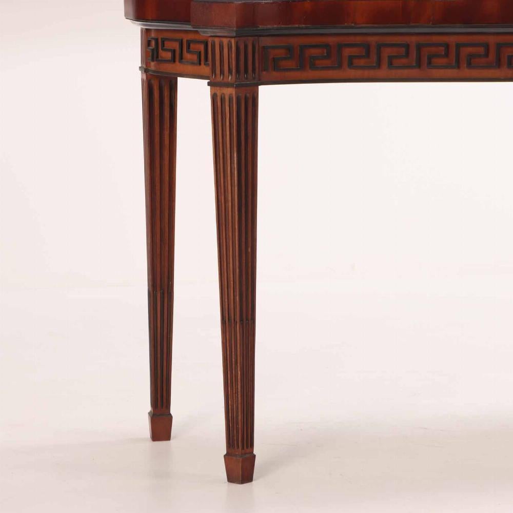 A Georgian-style console table with a fluted apron and legs embodies the refined elegance and classical influences characteristic of 18th-century British design.  Having a shaped and banded top as well as ebonized Greek key details, it is perfect