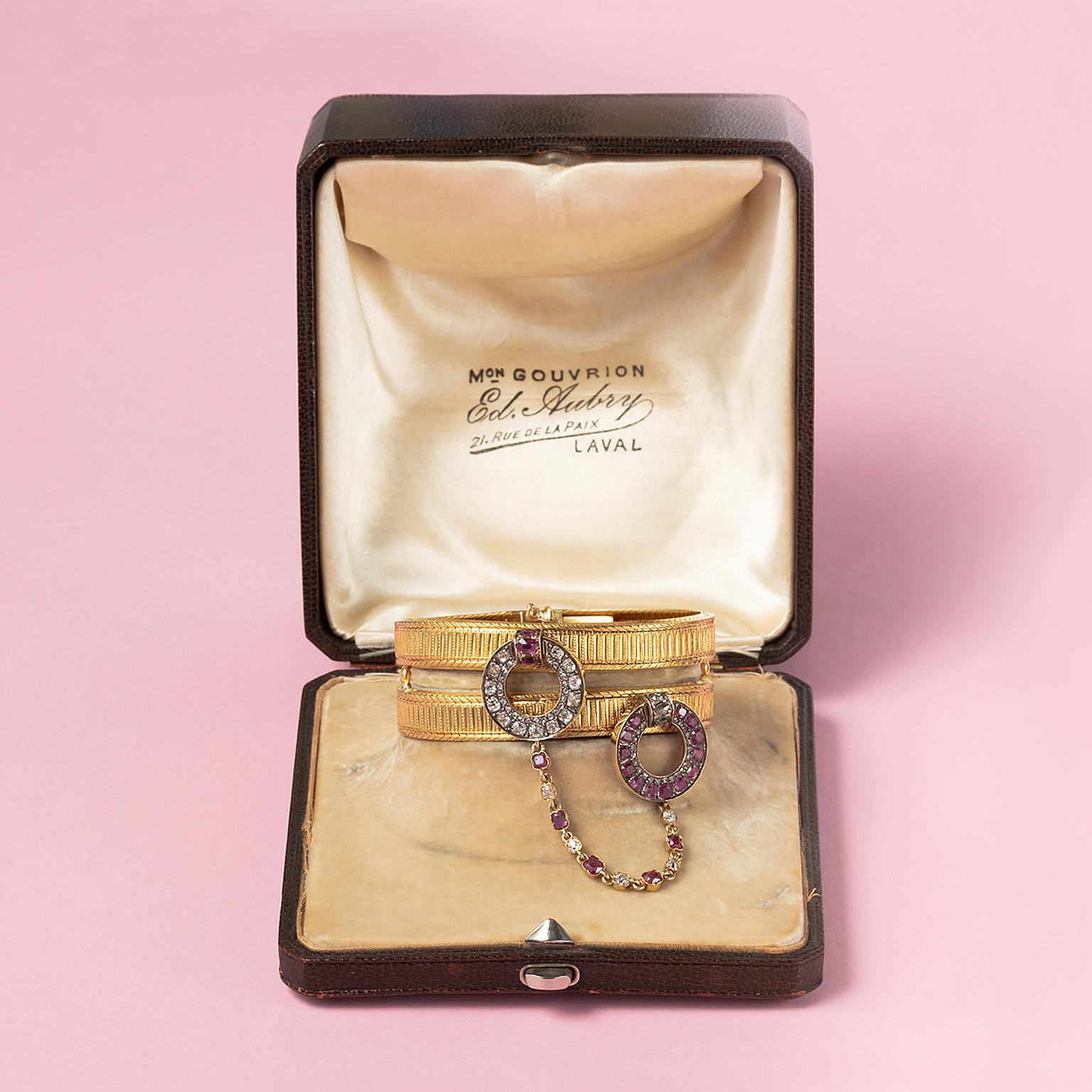 Ein Armband aus 18 Karat Gelbgold, bestehend aus zwei dicken, gerippten, flexiblen Armbändern, an denen jeweils ein Ring aufgehängt ist, von denen einer mit Diamanten im Kissenschliff und der andere mit Rubinen besetzt ist, beide mit einer