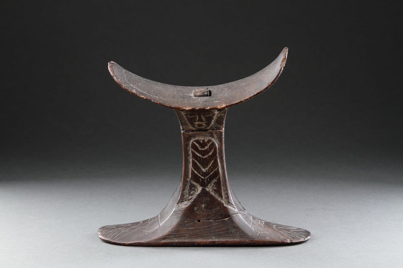 A Rare and Finely Carved Egyptian Wooden Headrest with a Carved Head to both Sides Representing the God ‘Bes’ (Protector of the Homestead) above Carved ‘Lilies’ Decorated to the Stem 

Fine old aged glossy patina 
An old paper label to the underside