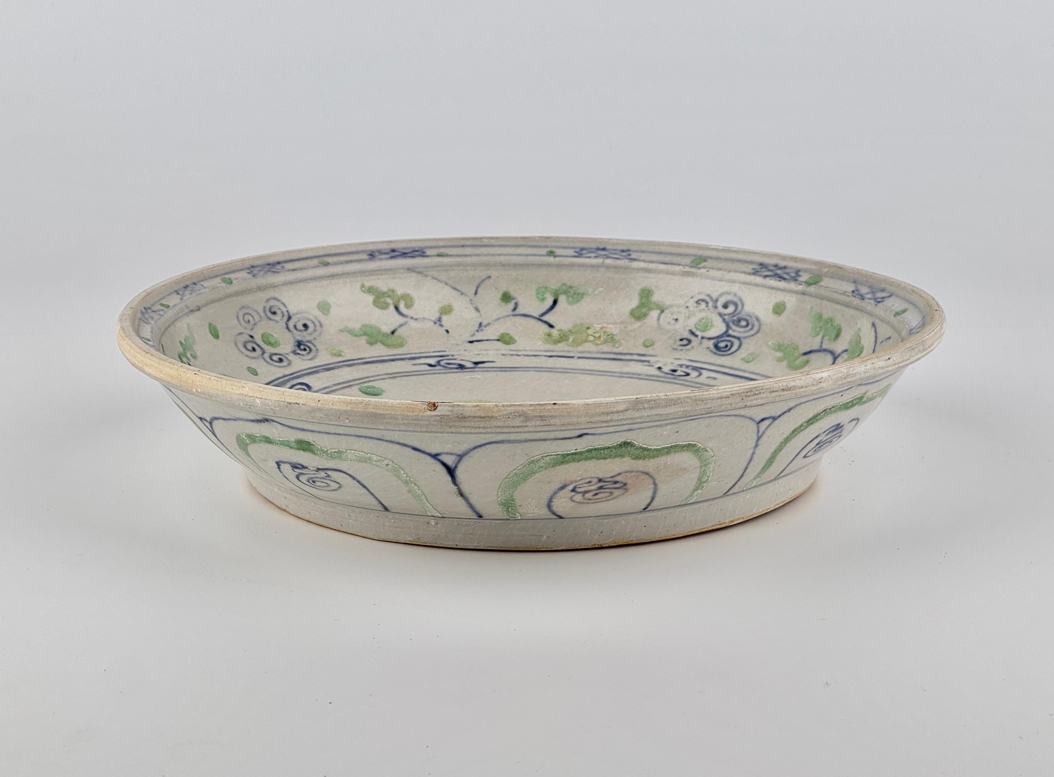 Cette assiette en céramique annamite présente un motif floral central réalisé en bleu sous glaçure, complété par de délicates feuilles peintes en vert - une caractéristique innovante des céramiques annamites de l'époque. Le bord est orné de motifs