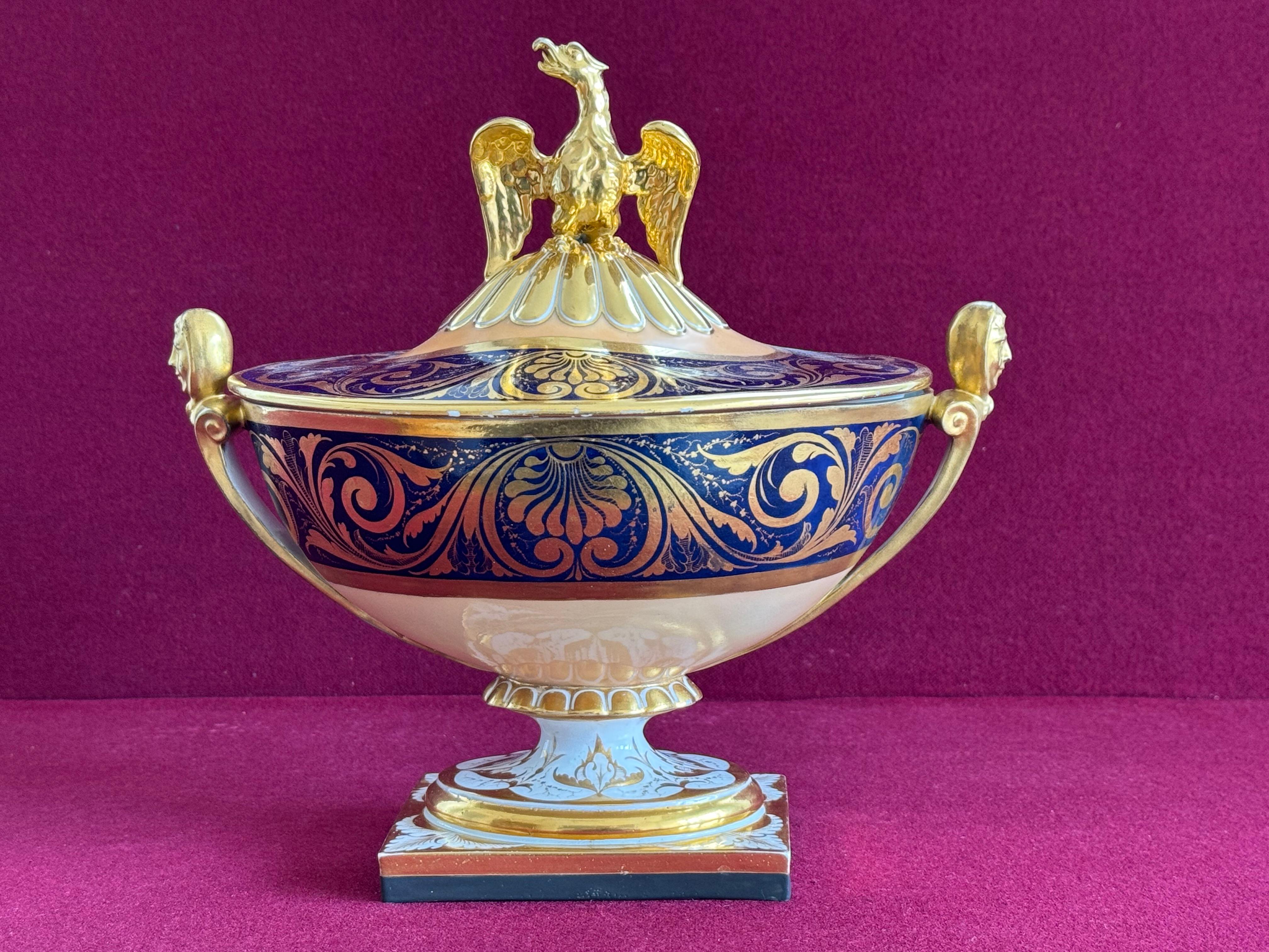 A rare and unusual form Barr, Flight and Barr Worcester vase and cover, circa 1810. Of low oval form, with mask head handles, the cover surmounted by an imposing eagle finial, painted with deep blue bands enlivened with neoclassical gilt borders, on